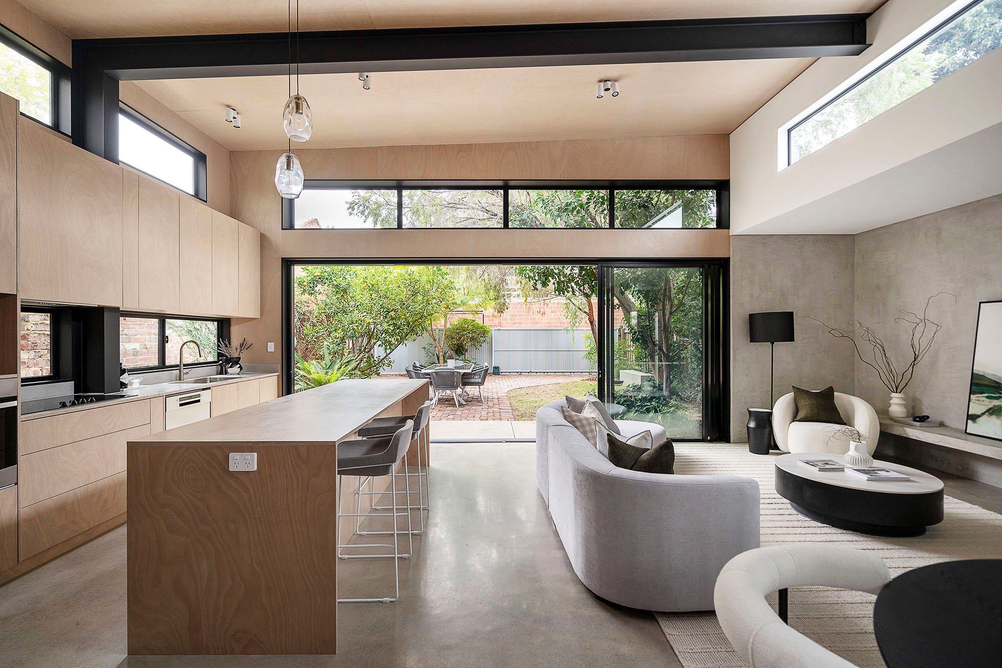 Open-concept modern living space with kitchen, bar seating, sofa, armchair, large sliding glass doors leading to backyard with patio furniture and greenery