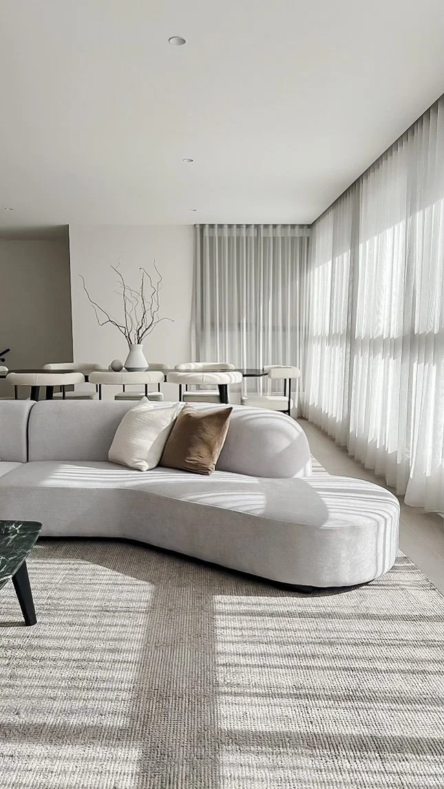 Modern living room with white sofa, beige and white pillows, large windows with sheer white curtains, and a dining table with white chairs in the background. Sunlight creates shadows on the light-colored rug.