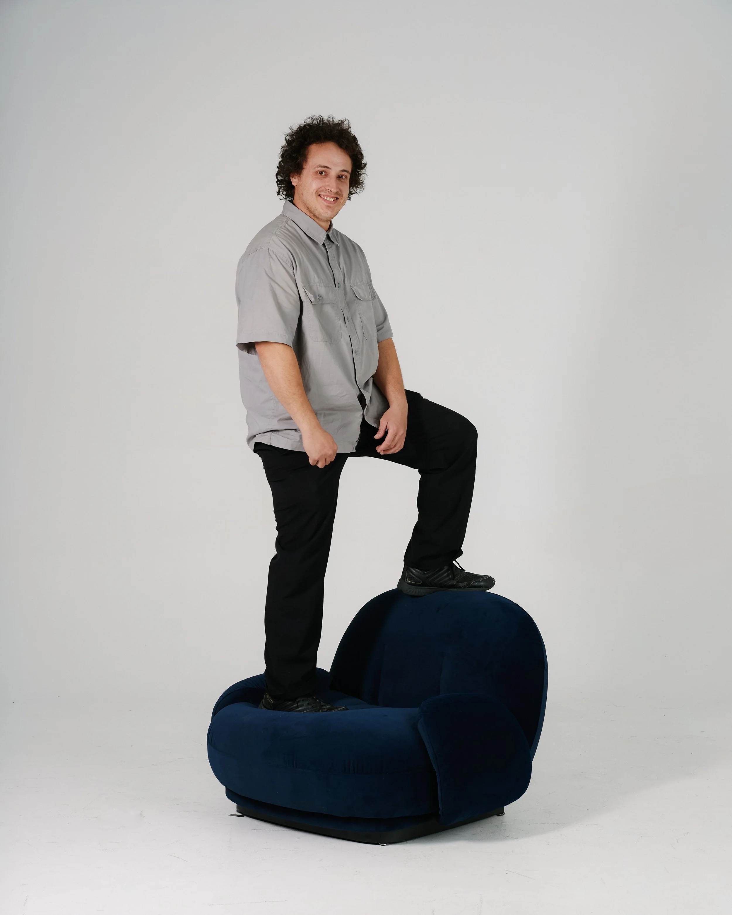 A bald man with a beard is sitting on a high stool, smiling, wearing a black t-shirt, black pants, and black shoes. His left arm has tattoos and he is wearing a watch and a ring.