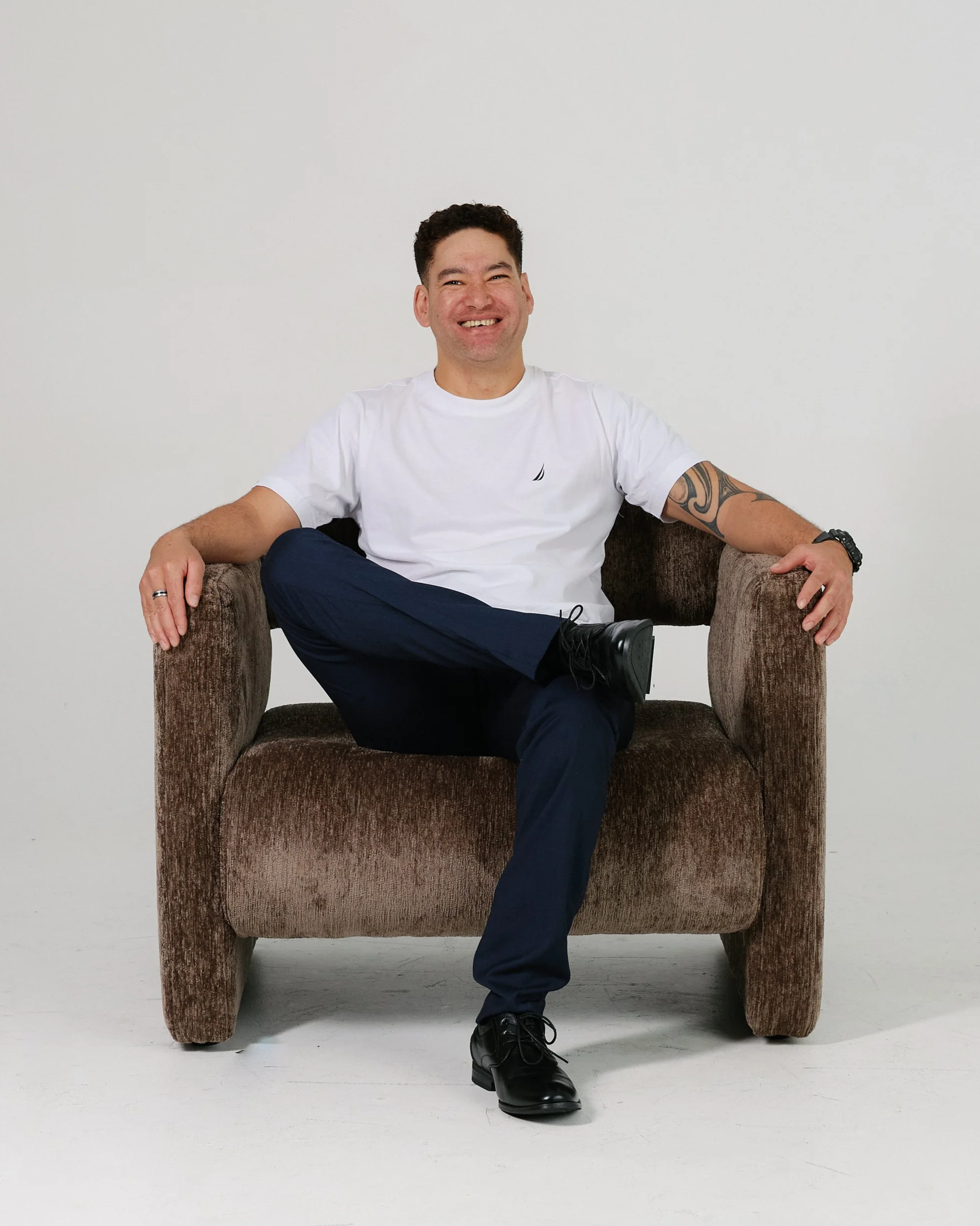 A man in black formal attire sitting on a textured, rounded chair against a plain, light-colored wall.
