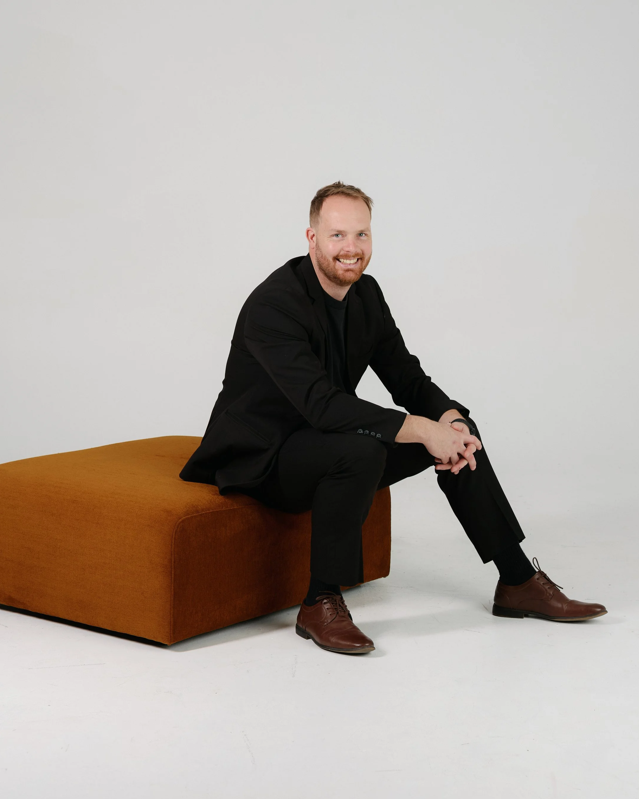 A man with red hair and beard, wearing a black blazer, black shirt, black pants, and brown shoes, sitting on a brown cushioned bench against a plain white background.