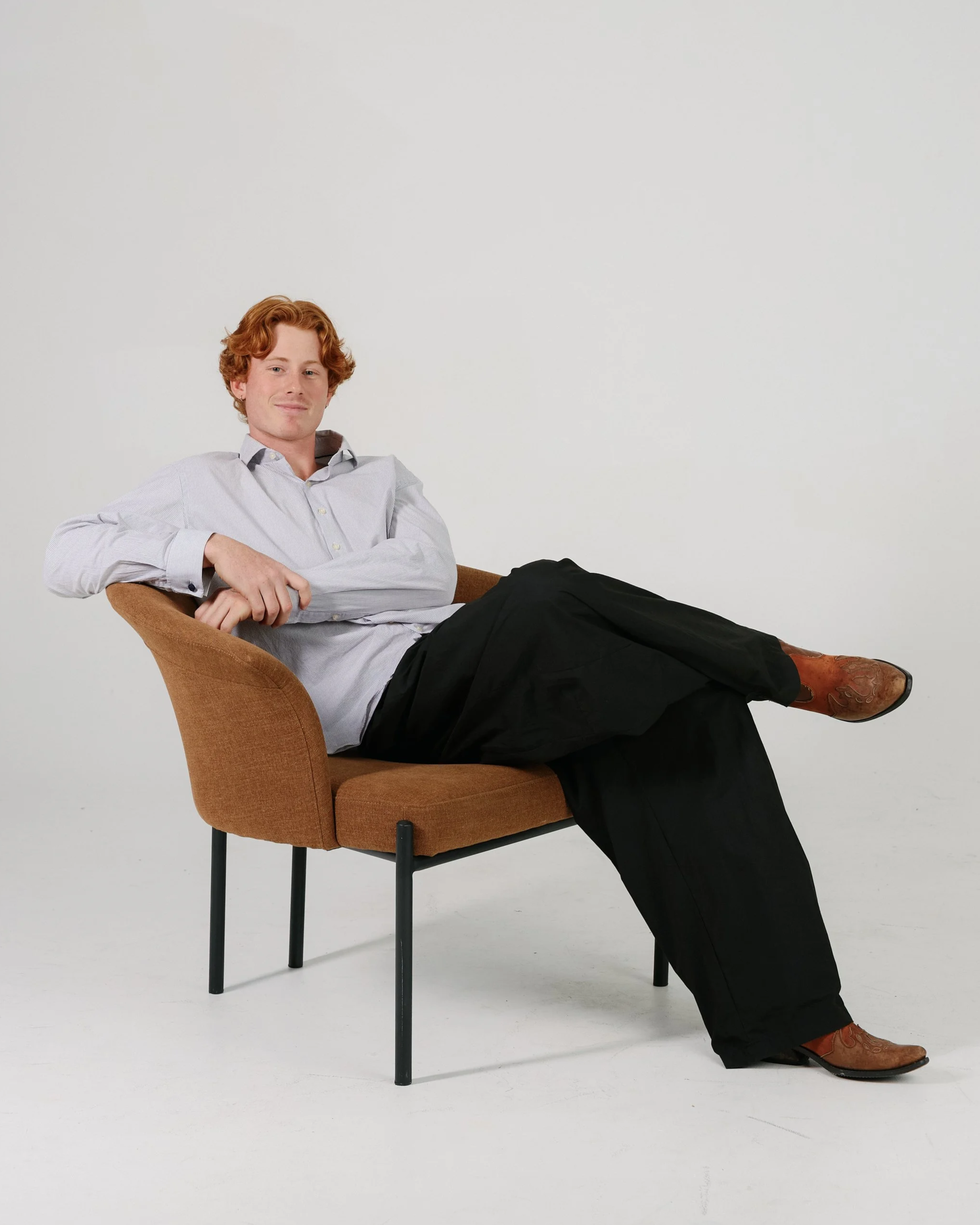 A young man with red hair, wearing a light-colored dress shirt and dark pants, reclining comfortably in a brown upholstered chair with black legs, against a plain white background.