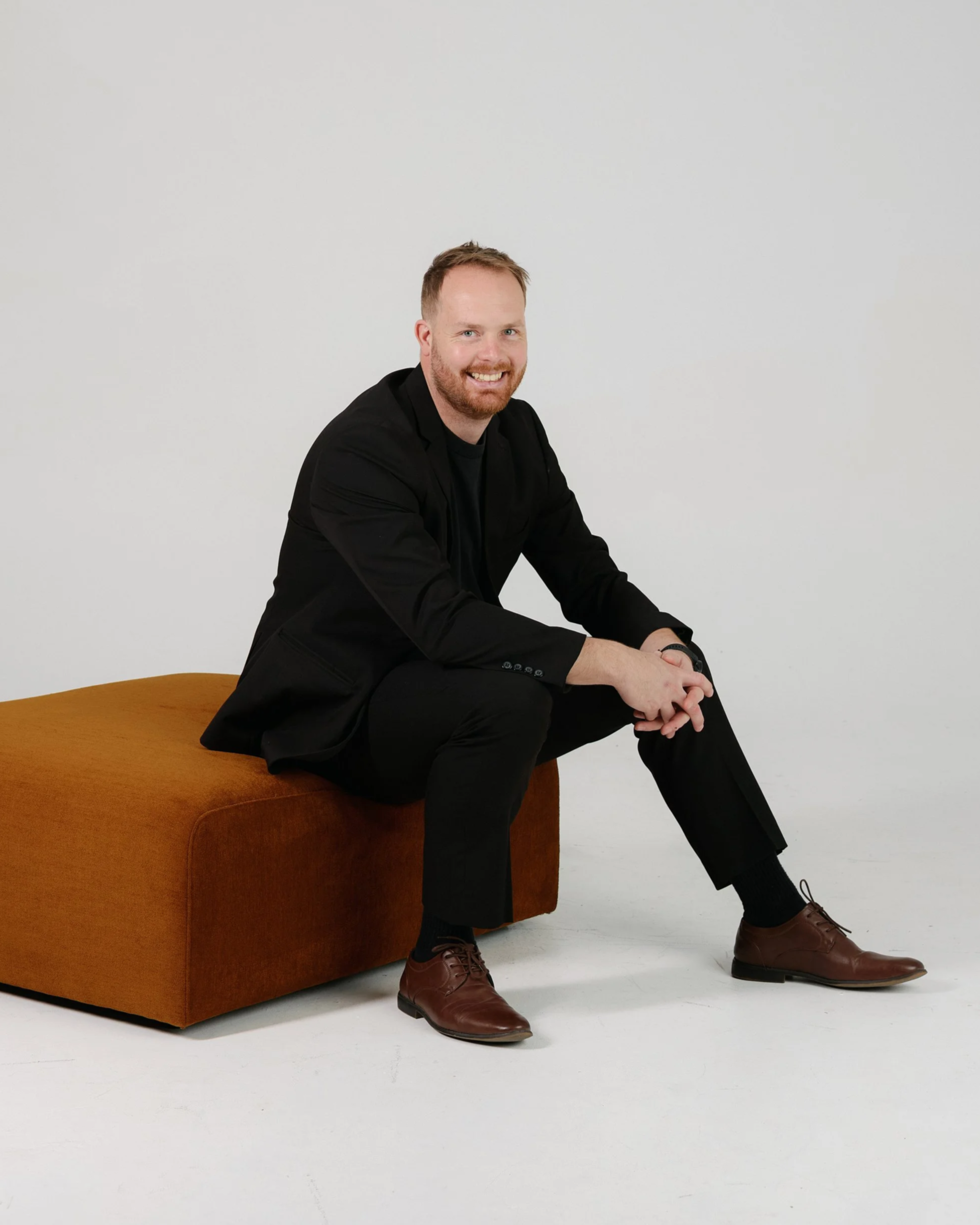 Black and white photo of a bearded man standing with his hands in his pockets, leaning against a tall stool in a plain room.