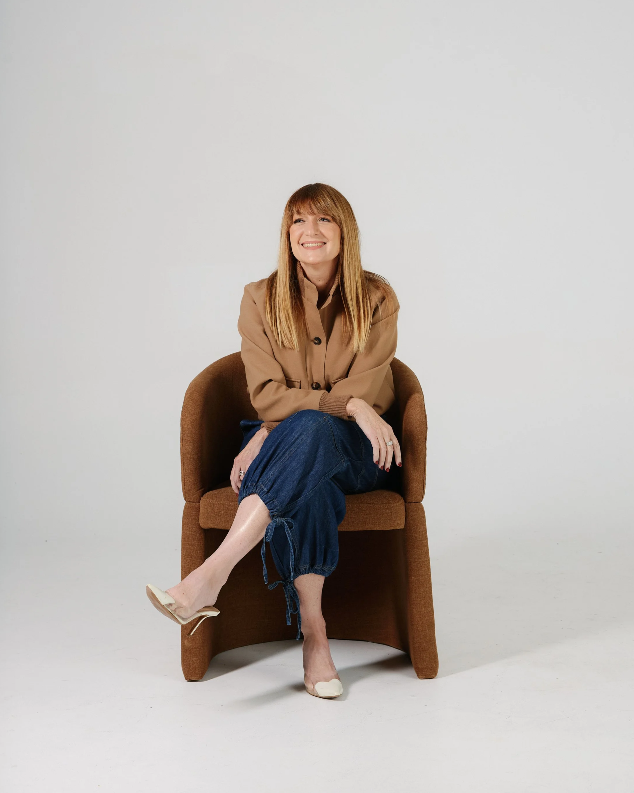 A woman sitting on a bench, smiling, with her right leg crossed over her left, wearing a black dress and high heels, against a plain background.