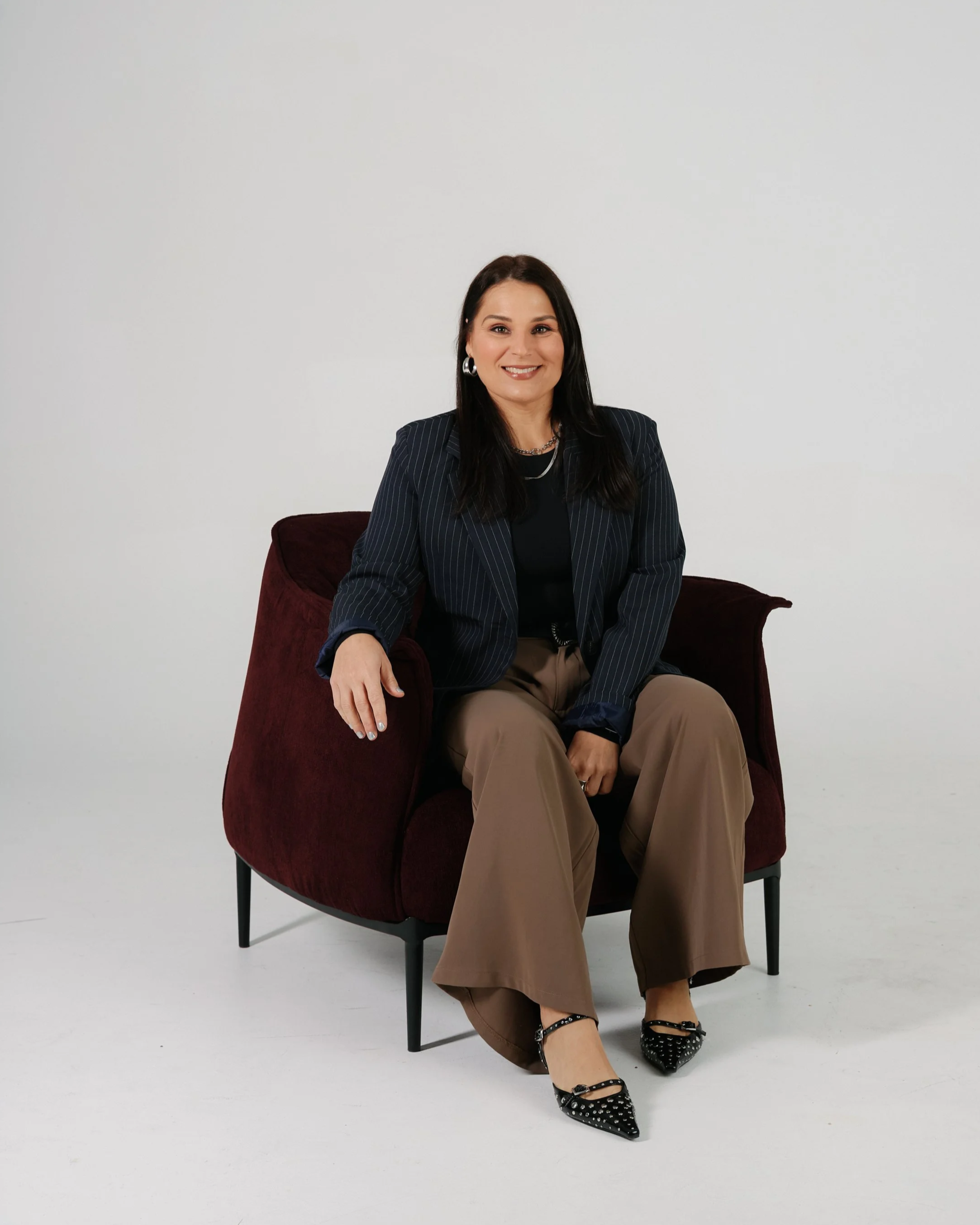 A woman sitting on a textured, ringed ottoman, wearing a dark outfit, high heels, jewelry, and a watch, smiling at the camera in a minimalist room.