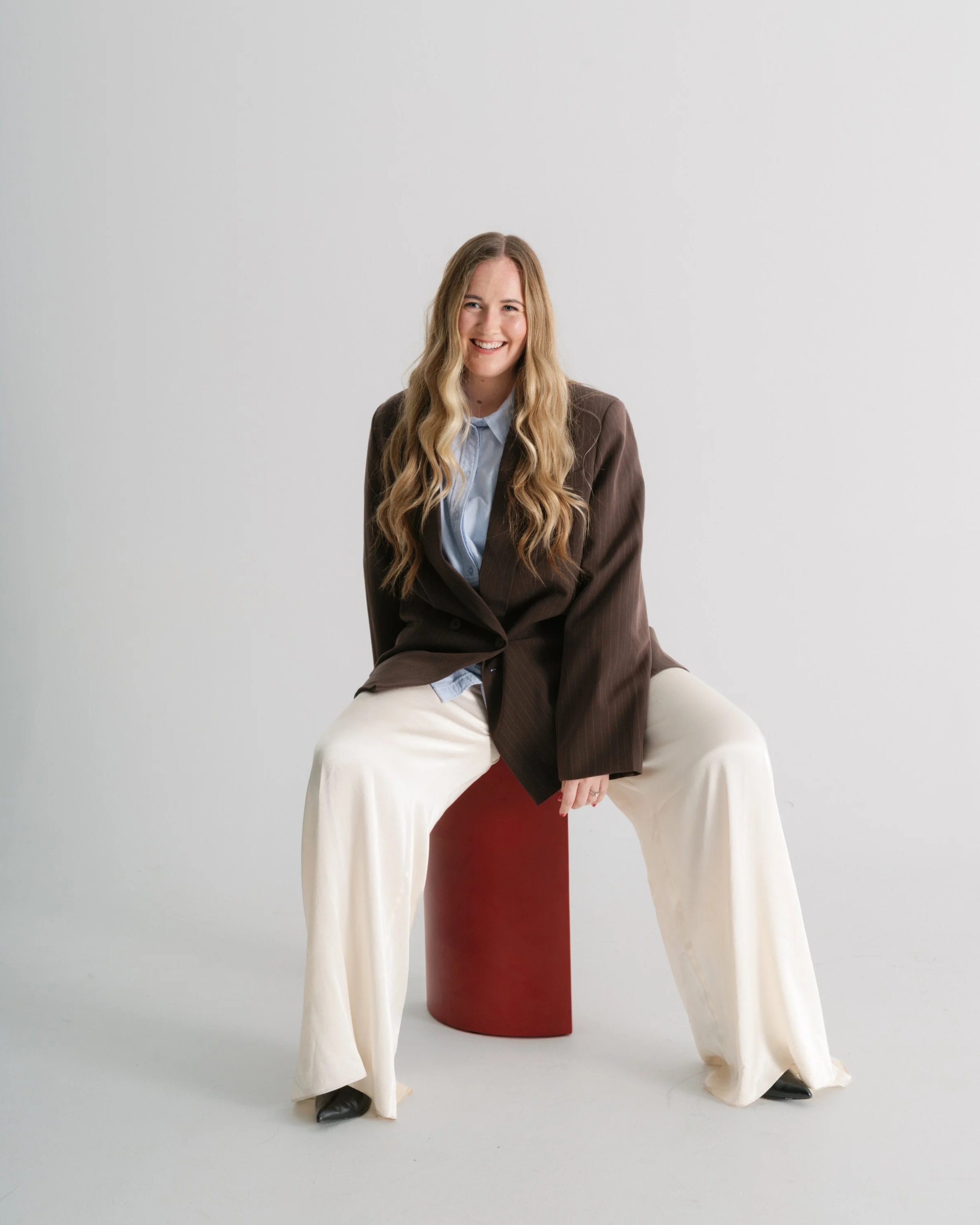 A woman with long wavy hair, wearing a dark outfit, seated casually on a light-colored armchair. She is holding a camera in her left hand, which is hanging down near the floor. The background is plain and minimalist.