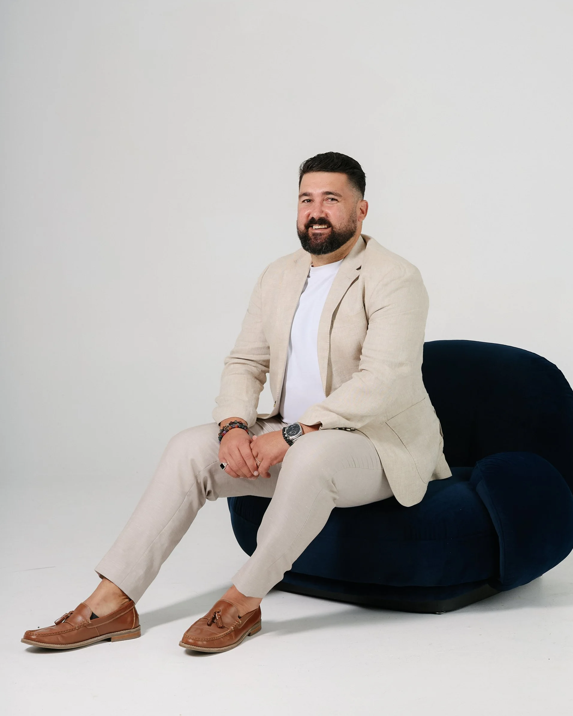 A man with dark hair, beard, and mustache, wearing a dark blazer, sitting in a light-colored chair against a plain white wall, with one leg crossed over the other.