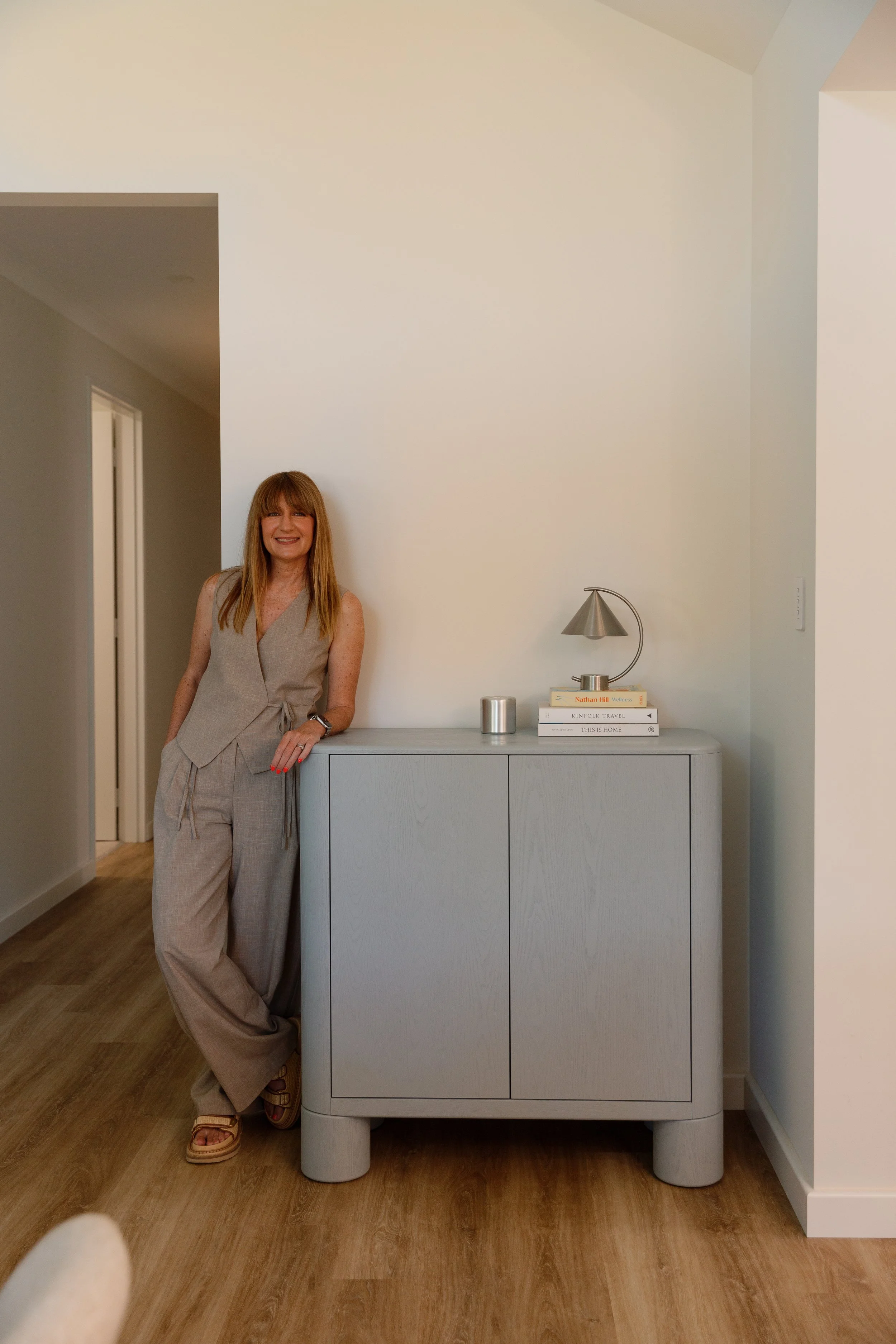 A woman with red hair and a beige jumpsuit standing and leaning against a white cabinet in a room with wooden flooring and white walls, smiling at the camera. On the cabinet, there are books, a modern table lamp, and a small metallic container. Part of the photographer's hand and a white object are visible in the bottom left corner.