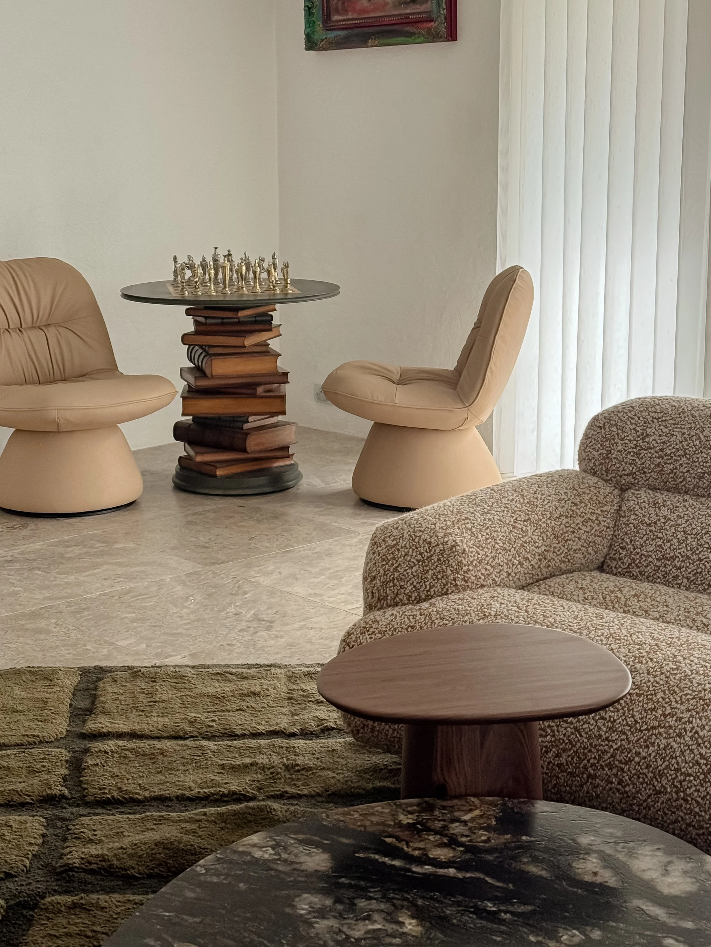 A living room with beige upholstered chairs, a round black marble-top table with a wood base, stacked books, and a chess set on top. The floor is marble, and there is a patterned rug partially visible. Vertical blinds cover a sliding door, and a colorful abstract painting hangs on the wall.