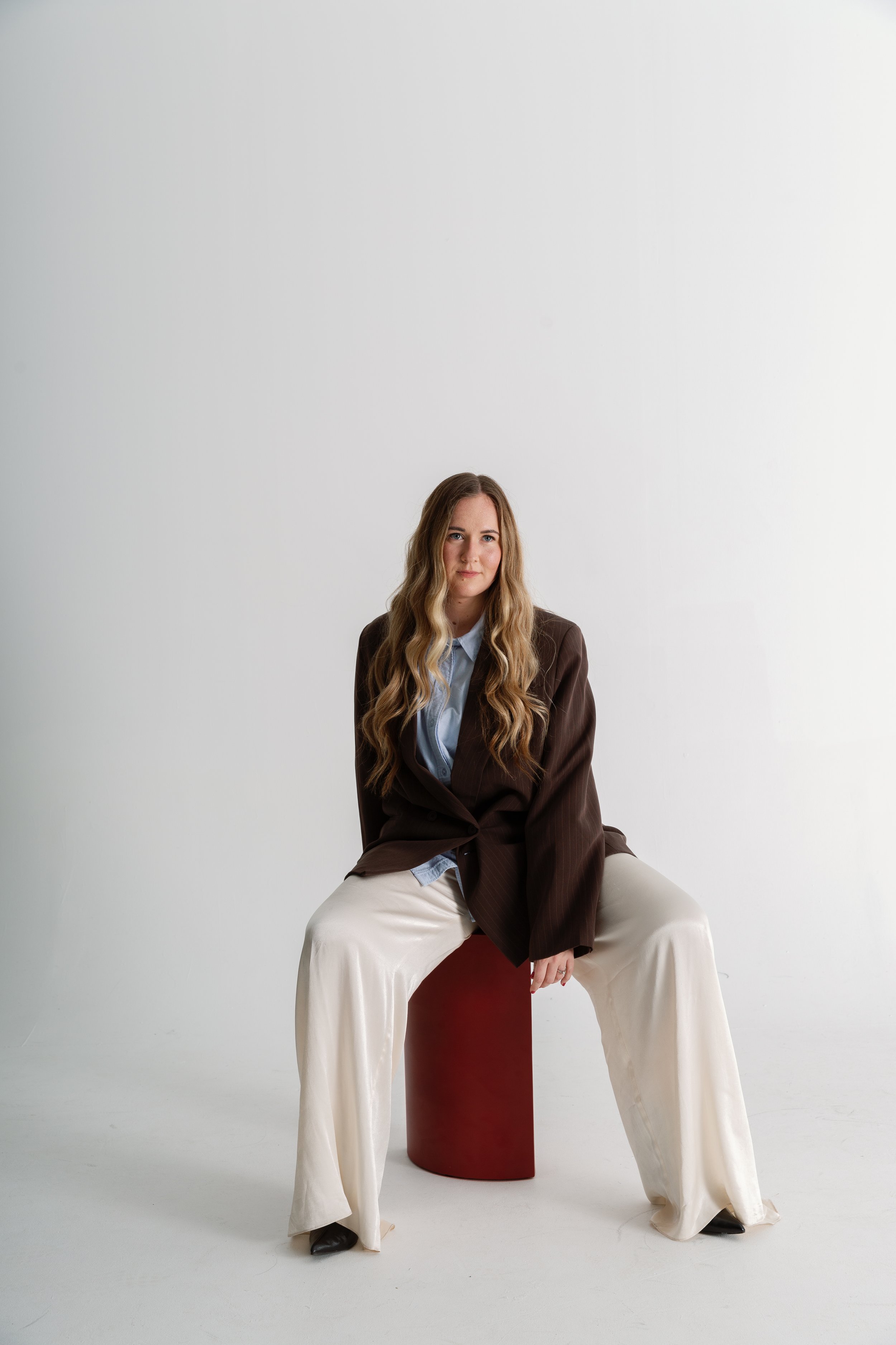 A woman with long wavy hair sitting on a red cube in front of a plain white background. She is wearing a brown blazer, light blue shirt, cream-colored wide-leg pants, and black shoes. She has a slight smile and looks directly at the camera.