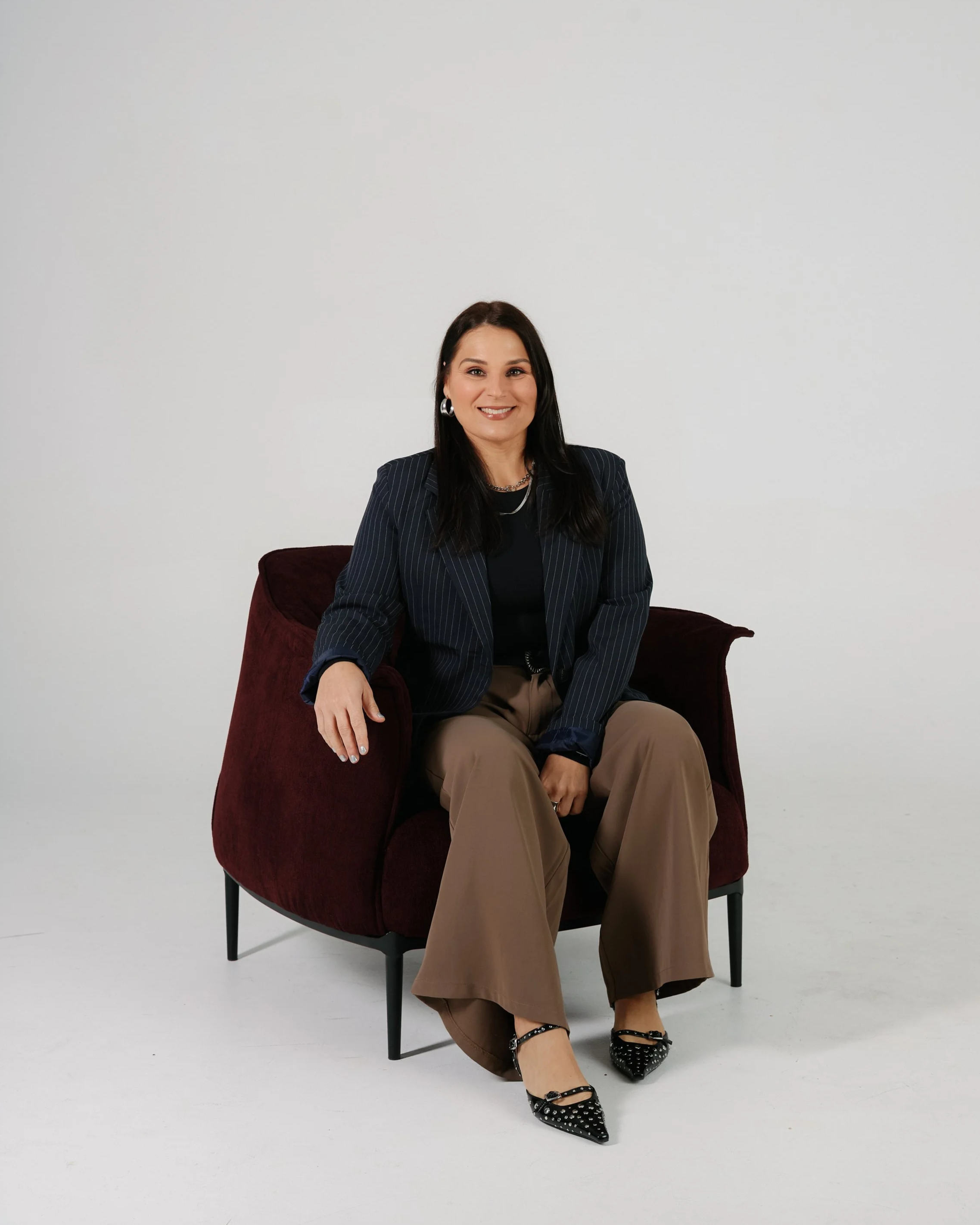 A woman with long dark hair, wearing a black top, pinstripe blazer, and beige pants, sitting on a burgundy armchair, smiling, against a plain white background.