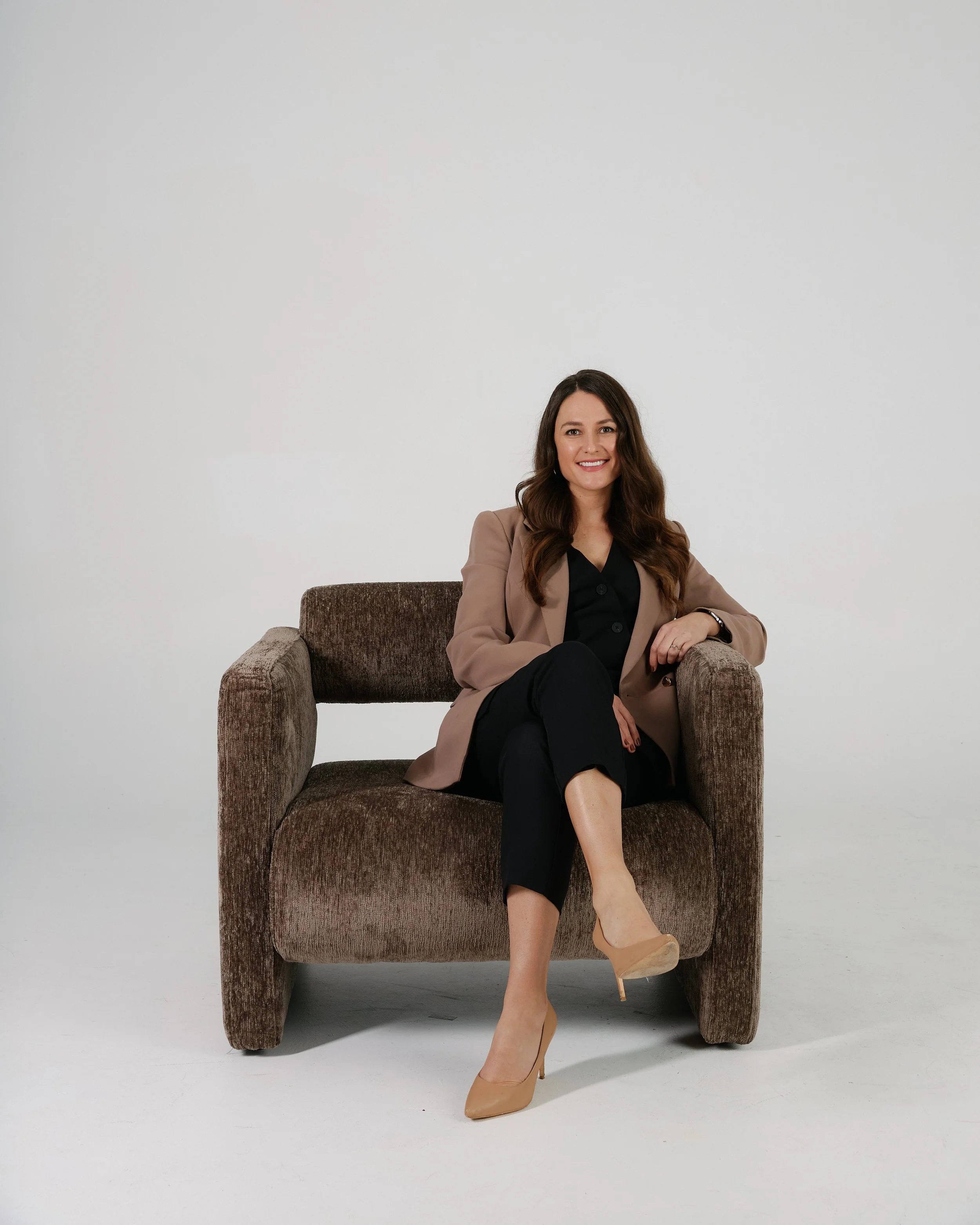 A woman sitting on a brown armchair wearing a beige blazer, black blouse, black pants, beige high heels, smiling against a plain white background.