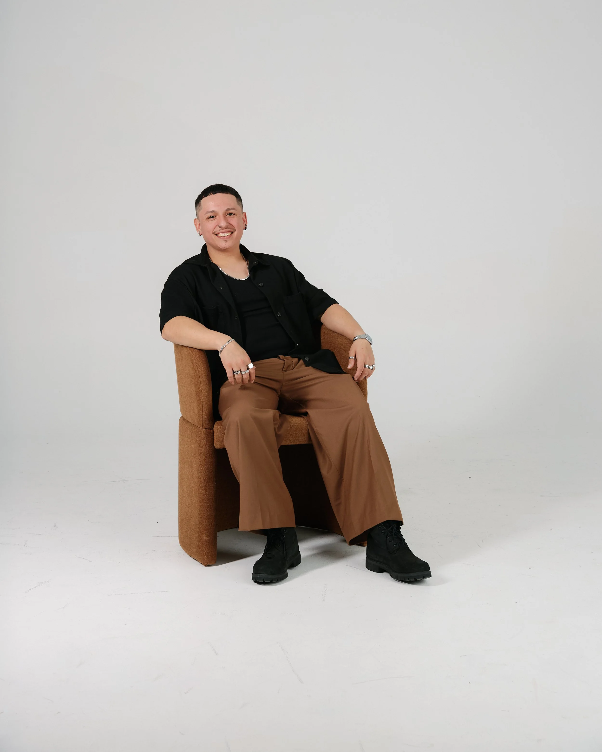 A young person with short dark hair, earrings, and a necklace, smiling while sitting on a brown chair in front of a plain white background. They wear a black shirt, brown pants, and black boots, with their arms resting on the chair's armrests.