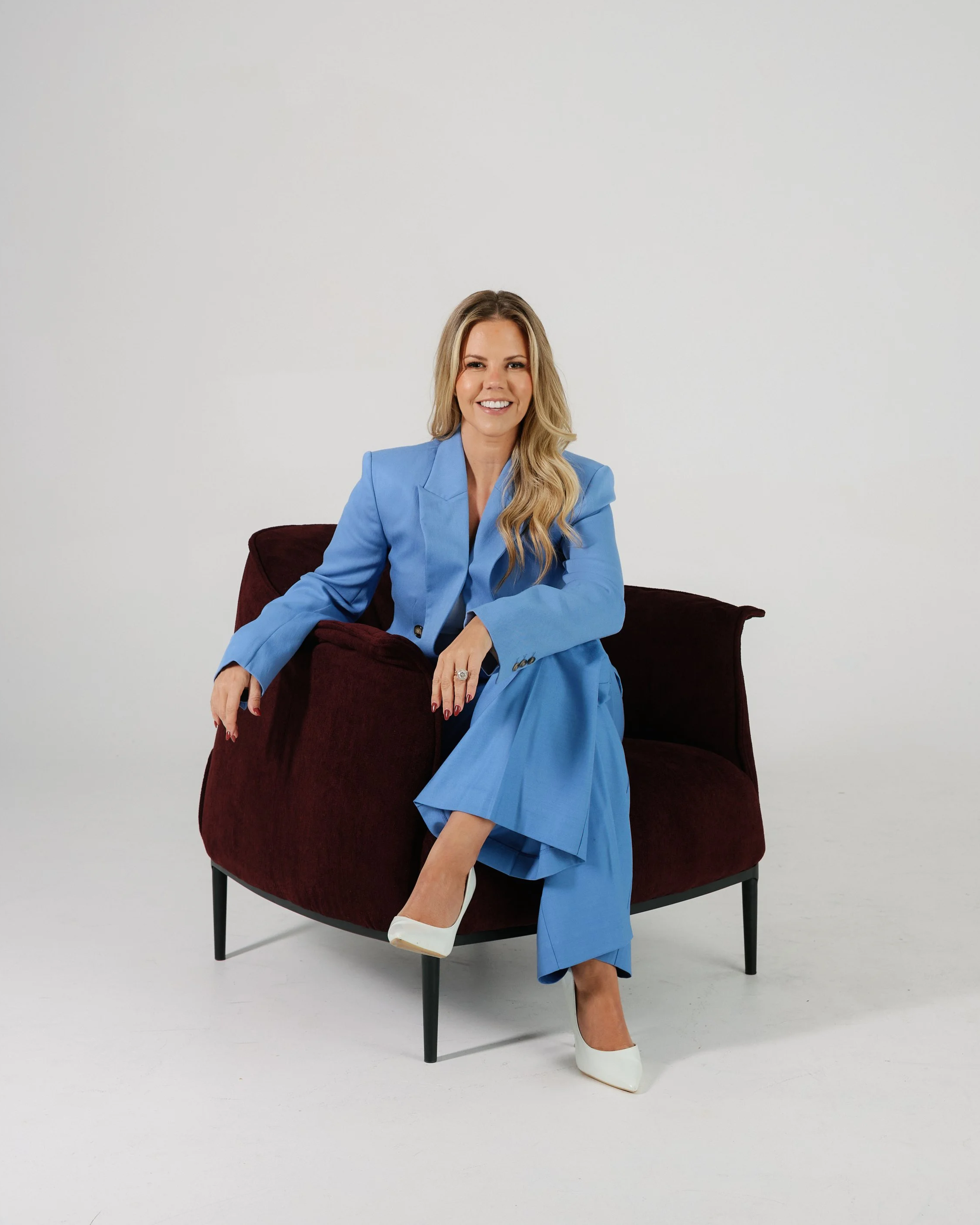 A woman with long blonde hair wearing a blue blazer and blue pants, sitting on a dark red armchair with black legs against a plain white background.