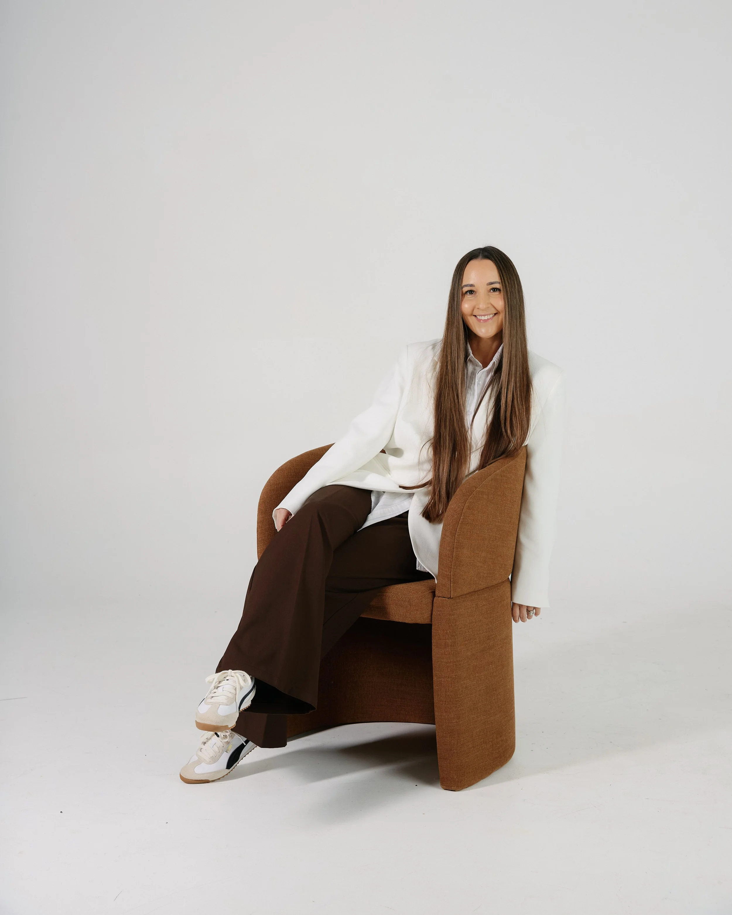 A young woman with long brown hair, white blazer, light-colored shirt, brown pants, and white sneakers, smiling and sitting sideways on a brown chair against a plain white background.