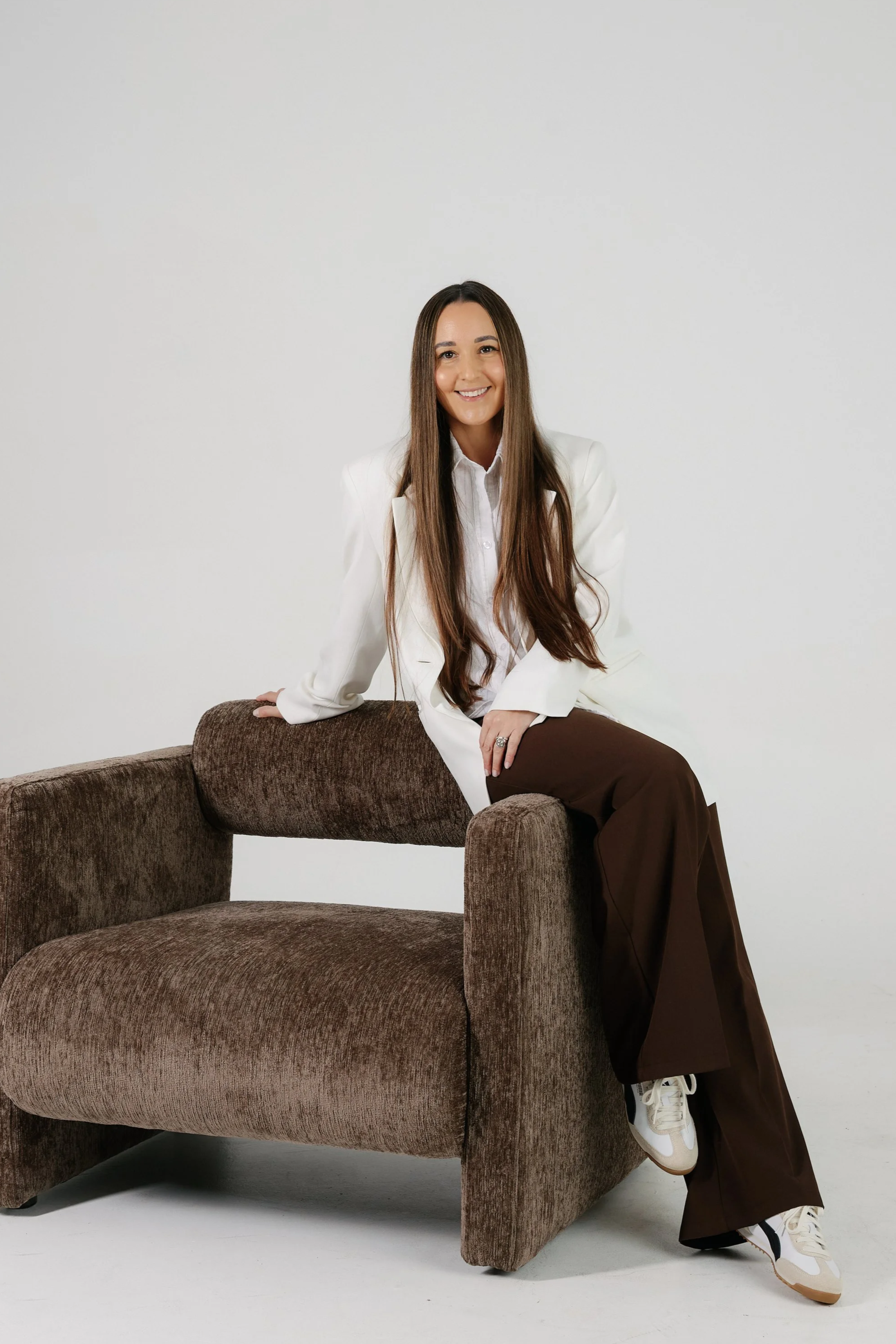 A woman with long dark hair, wearing a black blazer and pants, sitting on a tall stool and smiling.