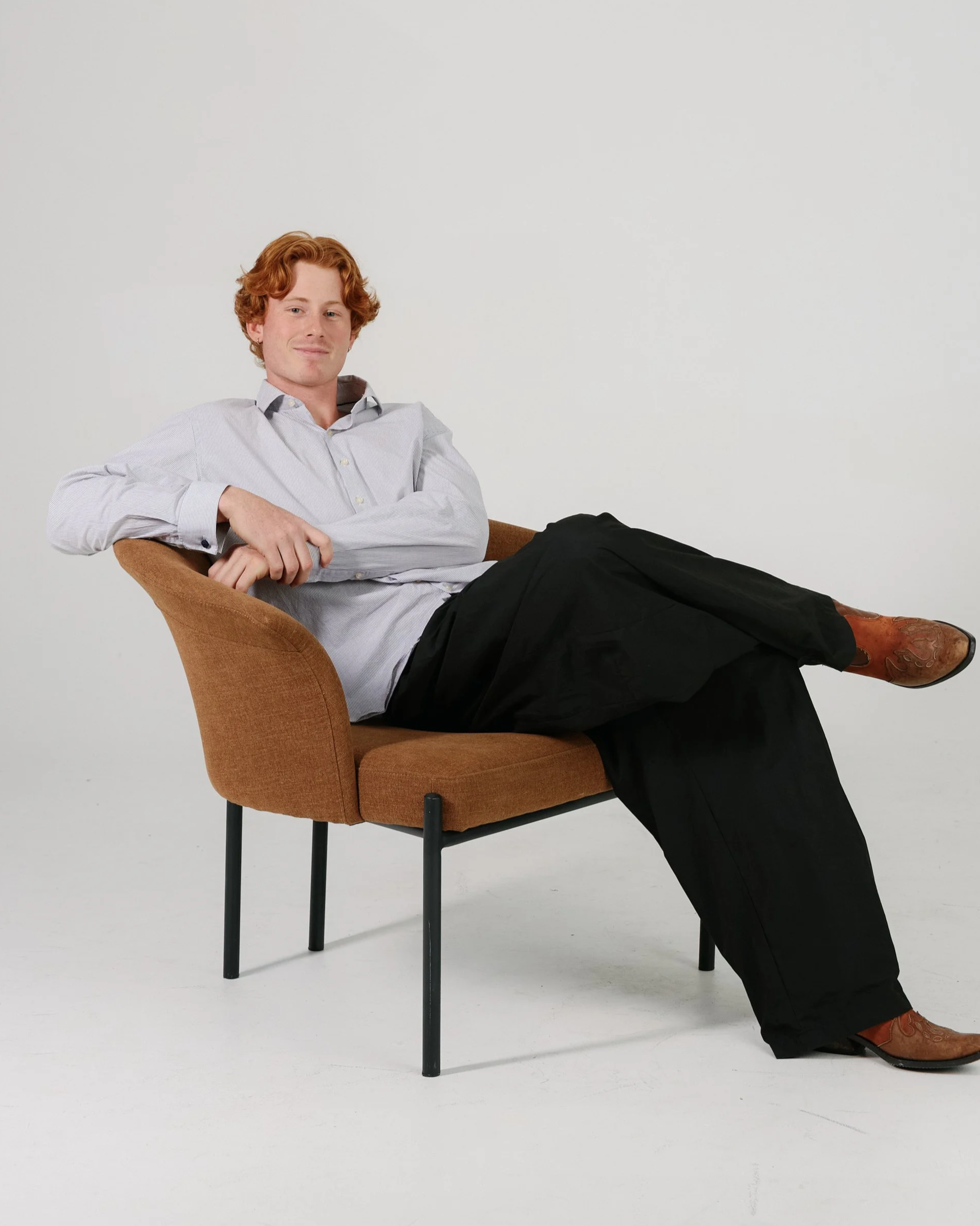 A young man with wavy hair leaning against a tall stool with a cushioned round seat, dressed in a black coat, black shirt, and black pants, standing in front of a plain wall.