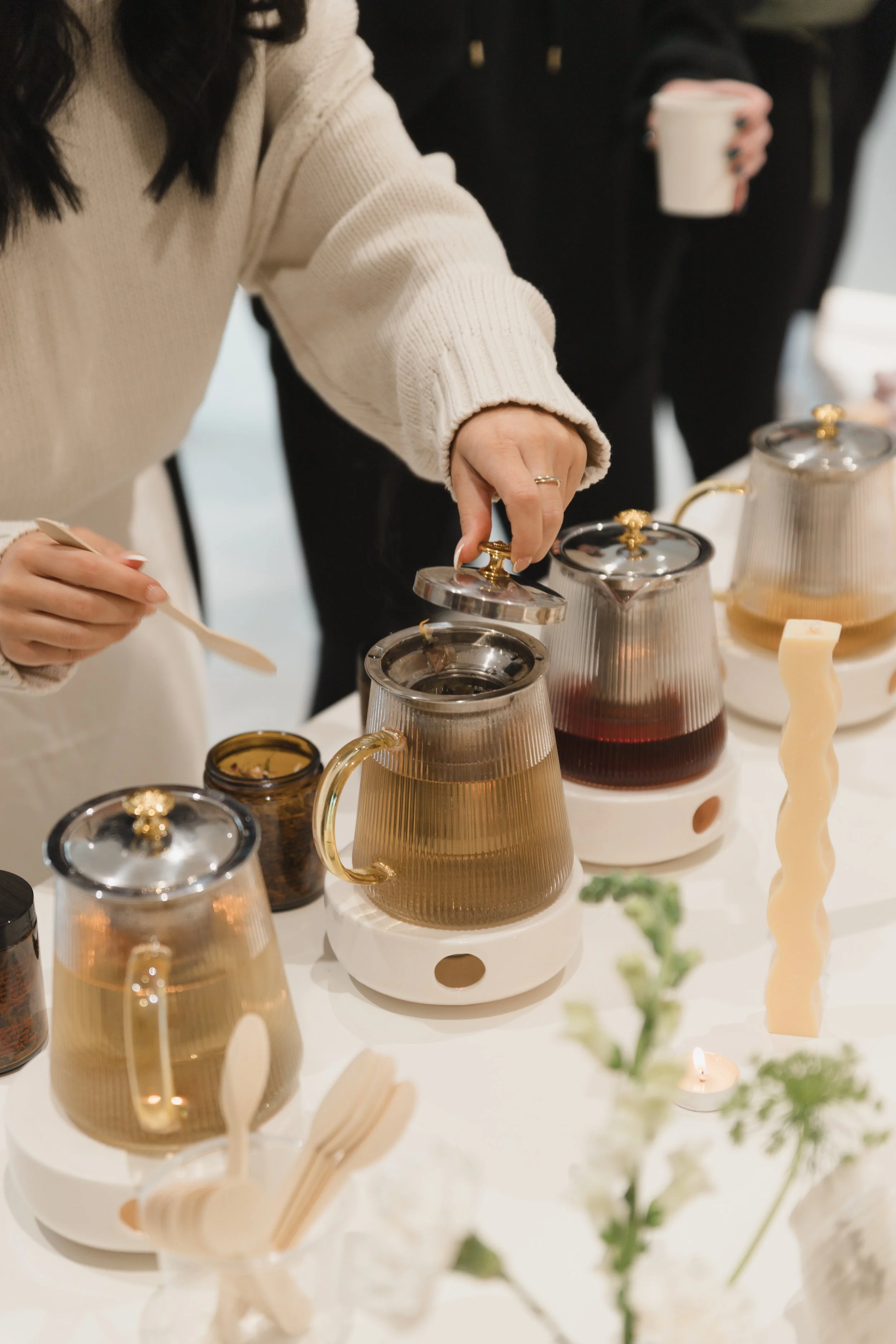 A person wearing a beige sweater reaching for a glass teapot with a black lid on a white display table, other teapots and jars are visible, and a person in black holding a cup in the background.
