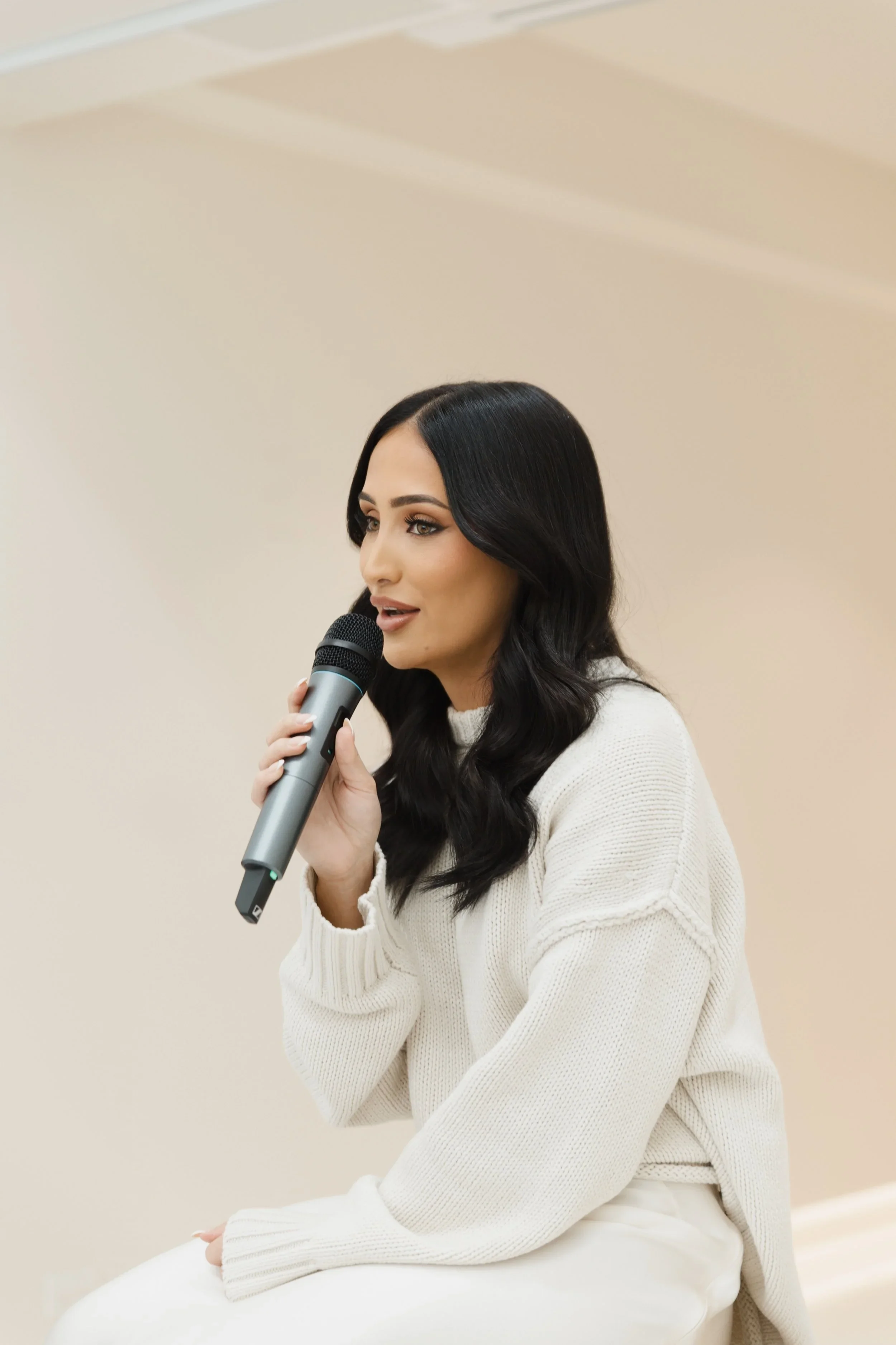 A woman with long black hair, wearing a cream-colored sweater, sitting and speaking into a microphone.