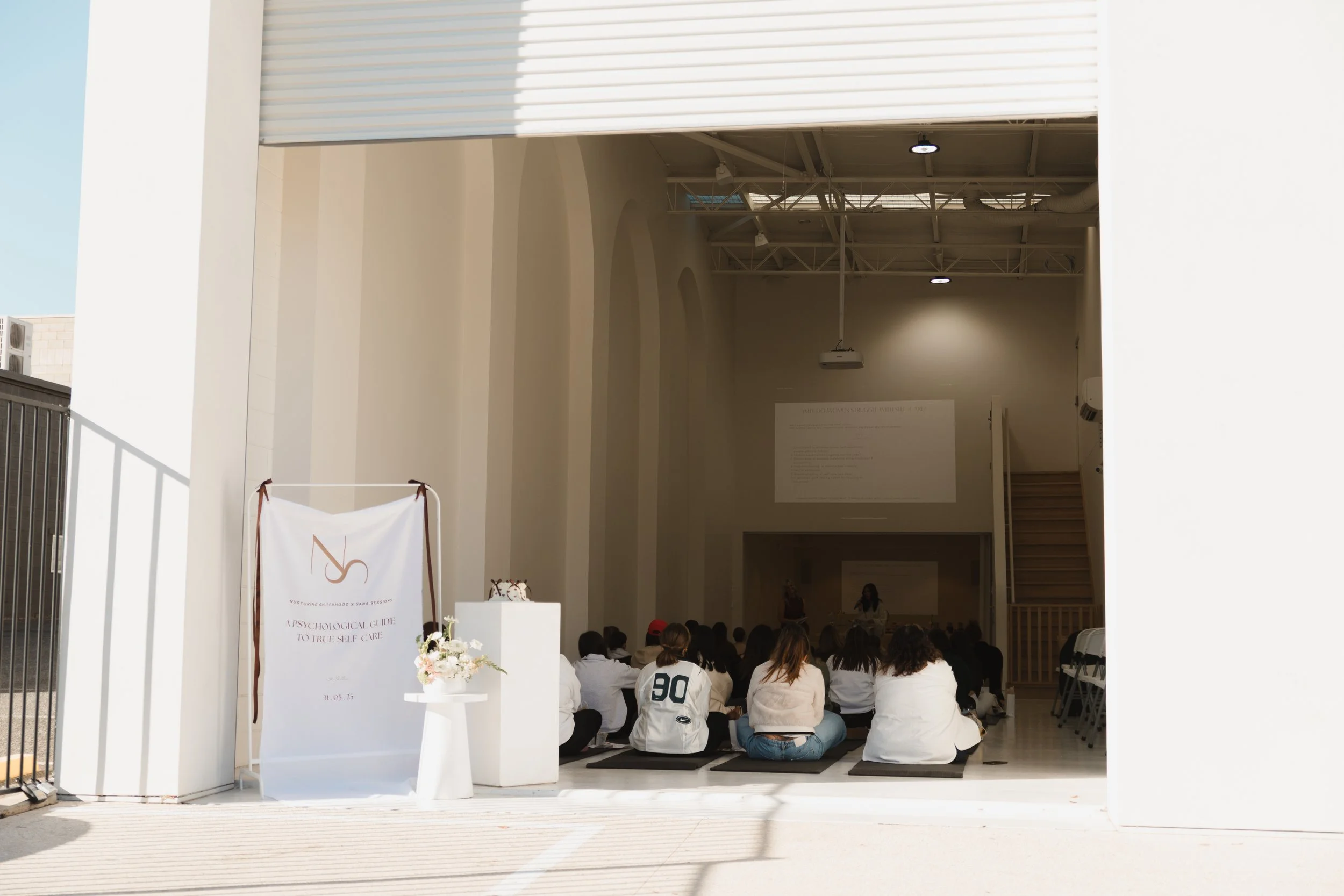 Group of people seated in rows listening to a presentation inside a large, open space with a high ceiling and natural light from windows above.