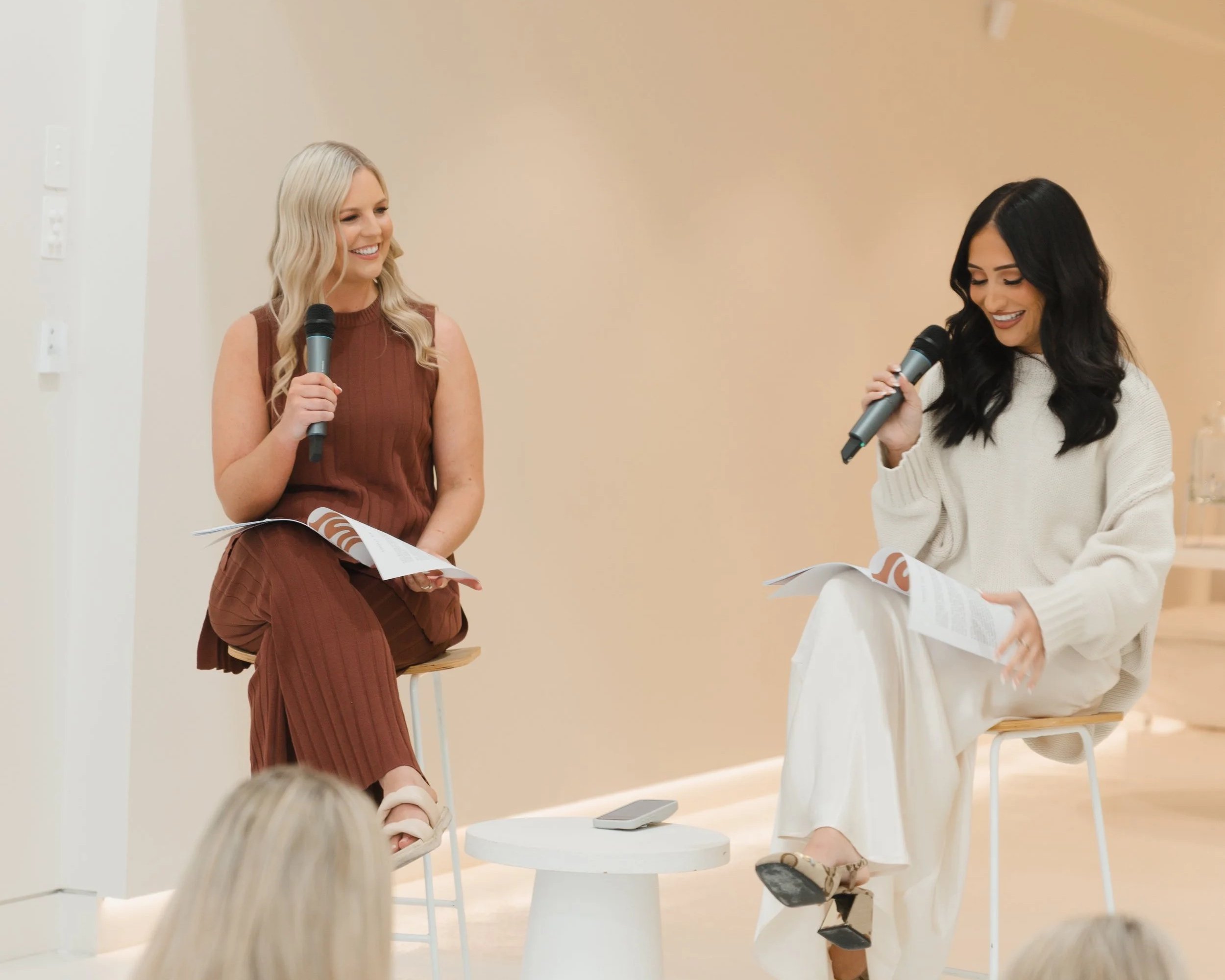 Two women holding microphones engaged in conversation, seated in chairs, with a small table and a smartphone on it, in an indoor setting with beige wall background.