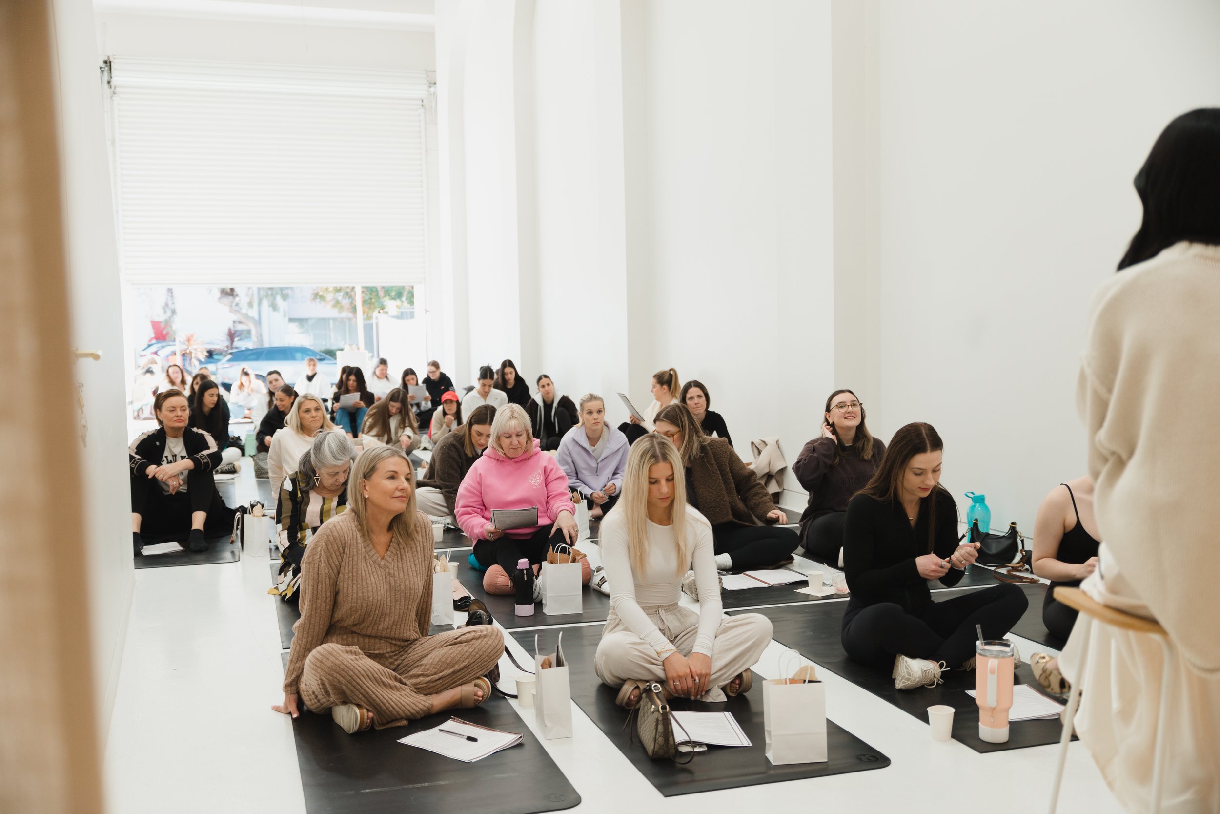 Group of people sitting on the floor in a spacious, bright room, participating in a workshop or class with notebooks and water bottles in front of them. A woman appears to be leading or speaking to the group from the right side.
