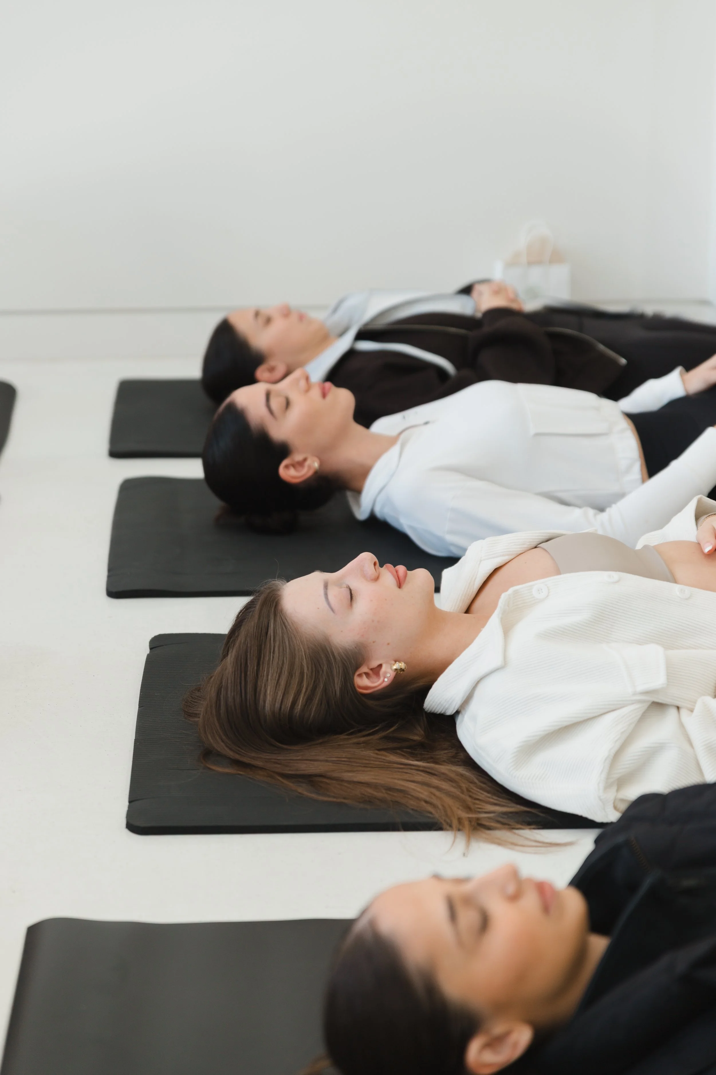 Four women lying on yoga mats with eyes closed, appearing to meditate in a bright white room.