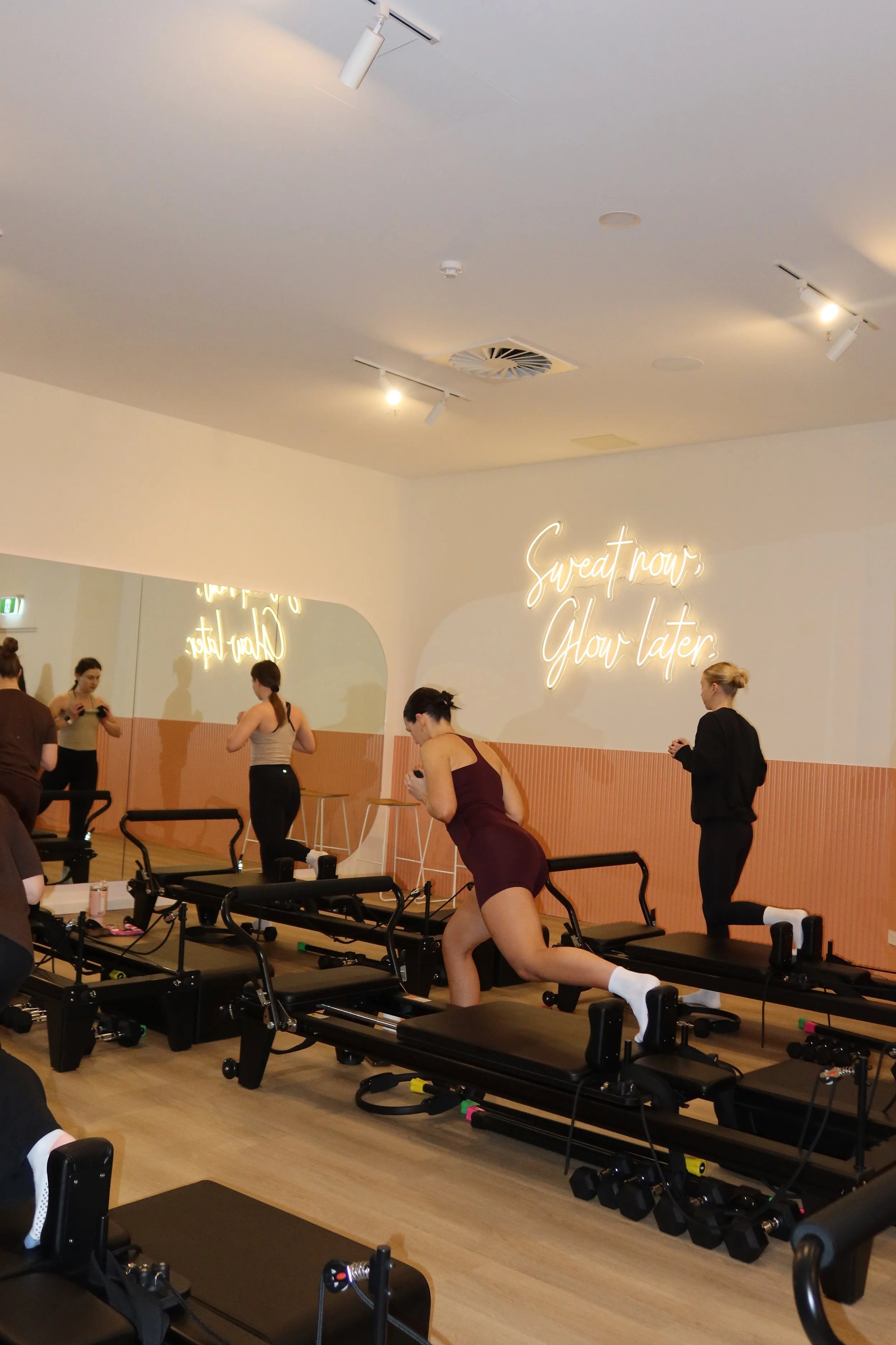 People engaging in a workout class at a fitness studio, with exercise equipment and a illuminated quote on the wall reading 'Sweat now, glow later'.