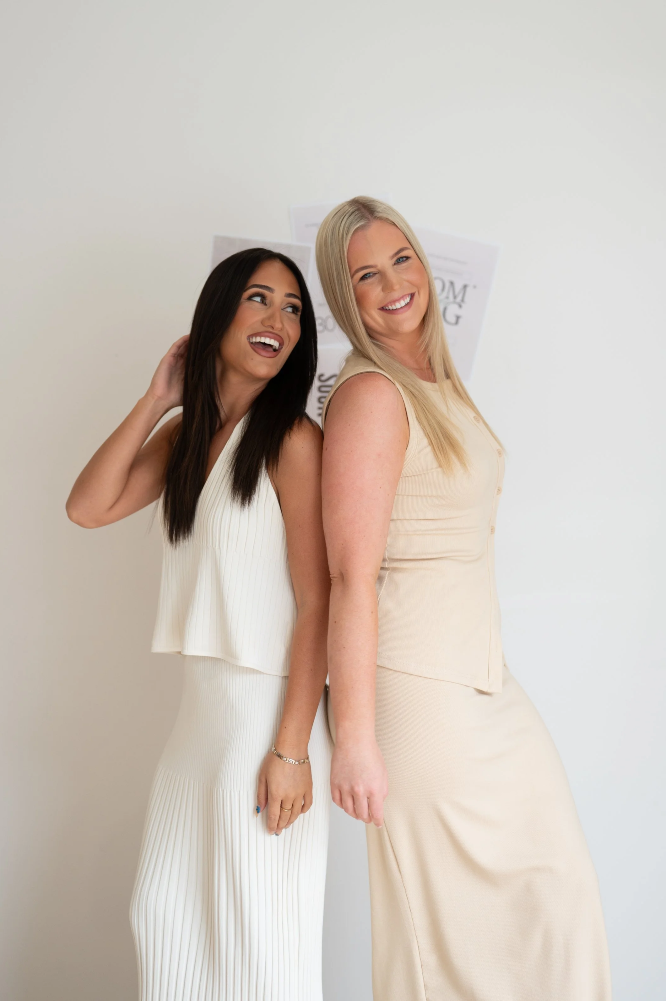 Two smiling women standing back-to-back, one in a white sleeveless top and pants, the other in a beige sleeveless dress, against a plain white background.