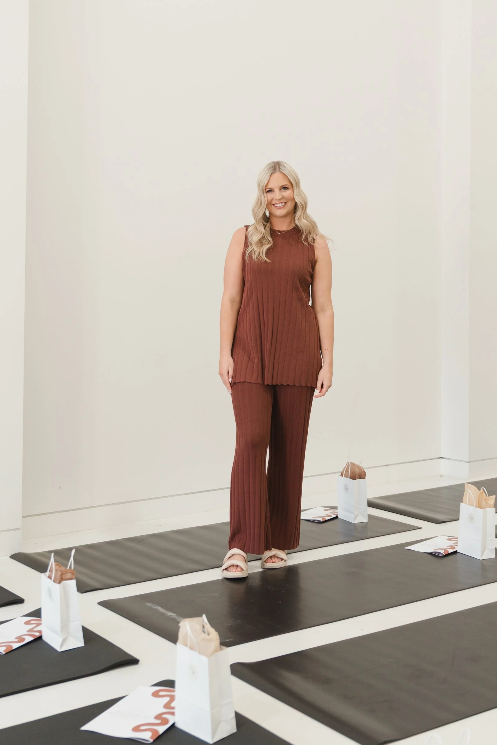 Woman standing on a yoga mat in a room with white walls, surrounded by small paper bags and magazines. She is smiling, wearing a sleeveless brown outfit and pink sandals.