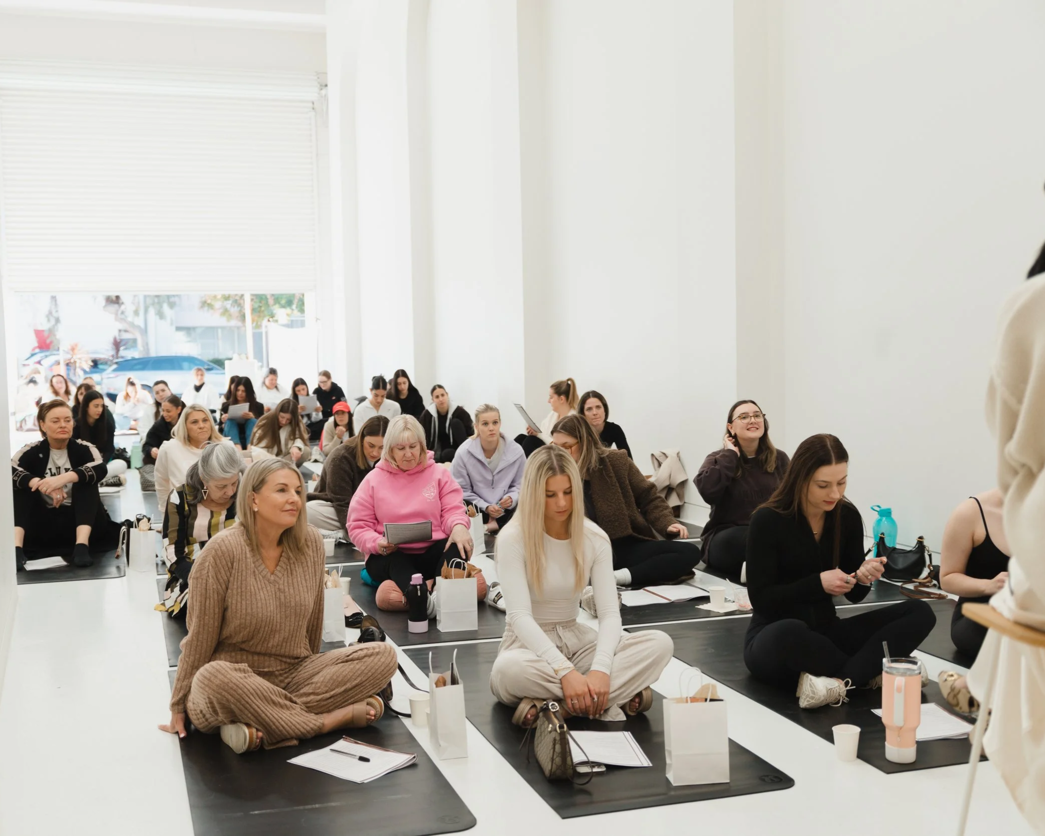 Group of people sitting on yoga mats in a bright indoor space, attending a workshop or class. Some are taking notes, others are listening attentively, with a few smiling, surrounded by personal belongings and bottles.