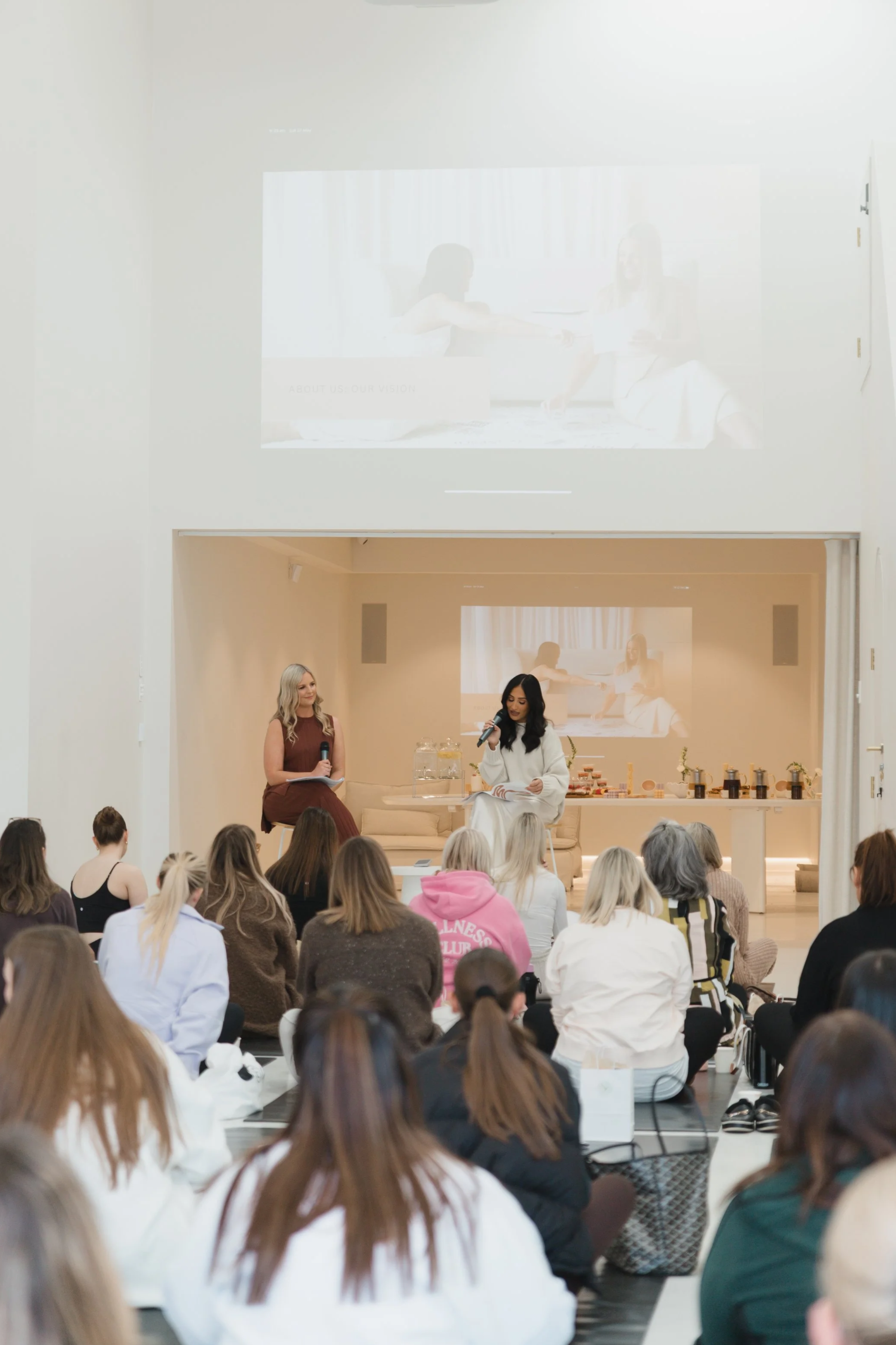 A large group of people attending a presentation or workshop in a bright, modern venue. Two women are seated on stage, one holding a microphone and the other with a notepad, while a large screen behind them displays a projected image of the same scene.