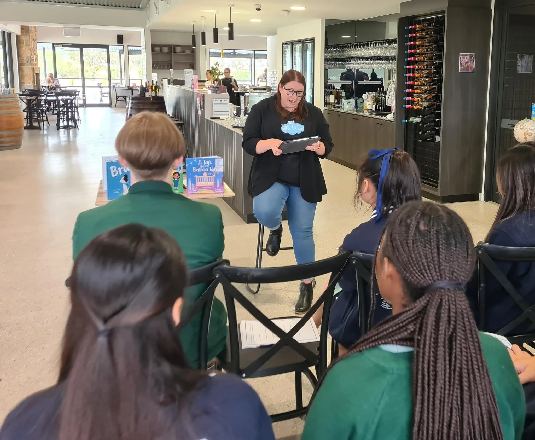 A group of children sitting at a table listening to a woman in a black blazer or jacket who is standing and holding a tablet or notepad. The setting appears to be a cafe or restaurant with a bar area in the background, wine bottles stored in a wine rack, and books displayed on the table.
