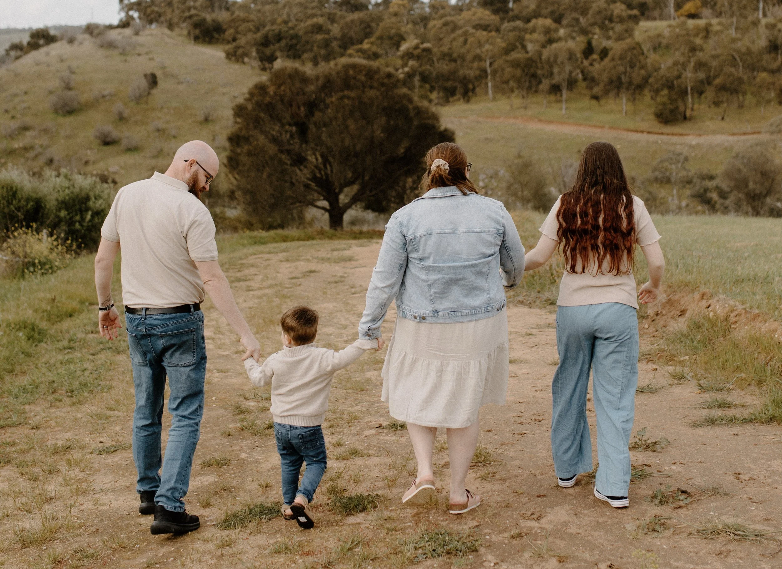 A family of five walking outdoors on a dirt trail through a green, hilly area with trees and shrubs, during daytime.