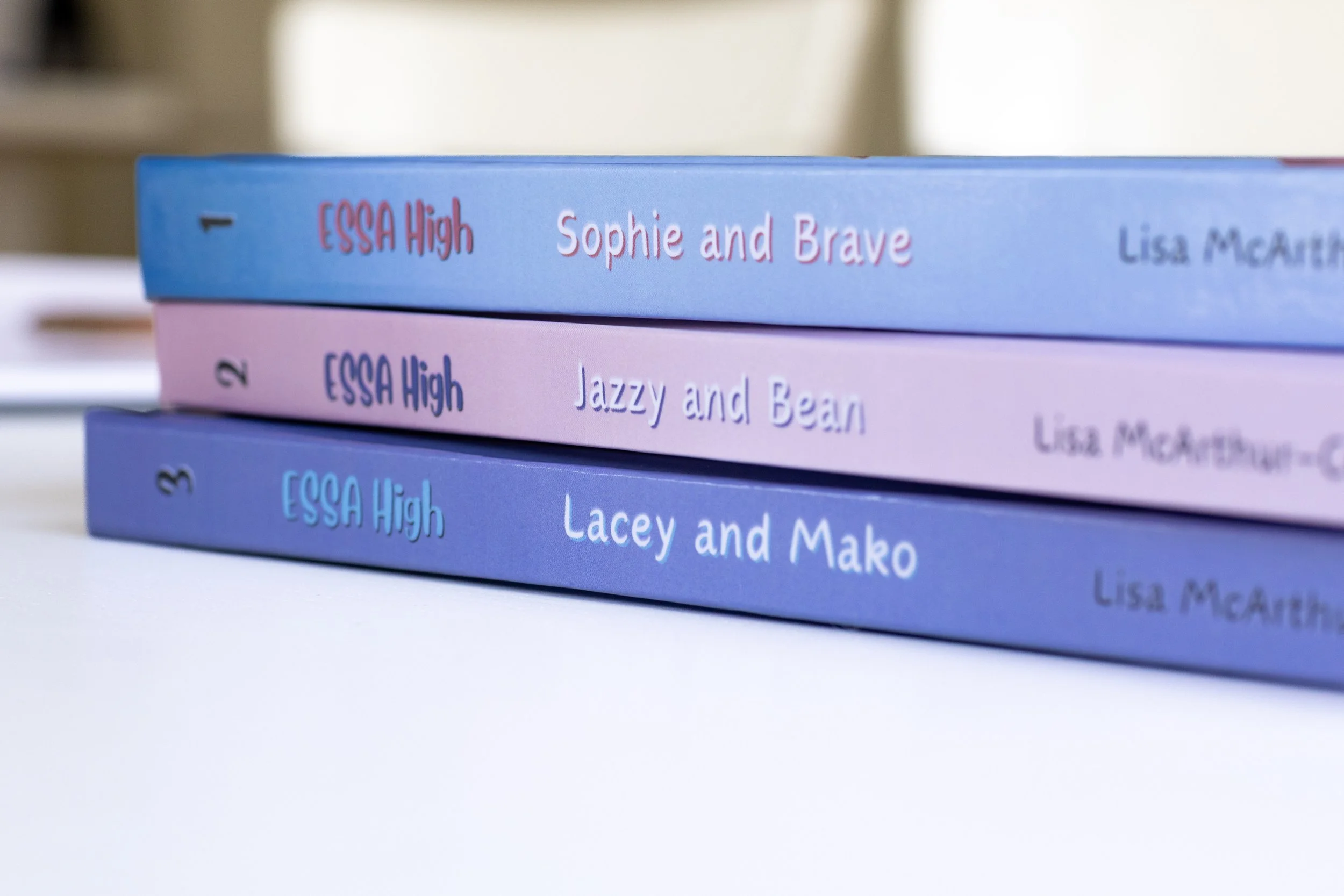 Close-up of three stacked books with colorful covers. The top book has a blue cover titled 'Sophie and Brave' by Lisa McCarthy, the middle book has a pink cover titled 'Jazzy and Bear' by Lisa McCarthy, and the bottom book has a purple cover with an incomplete title and author.