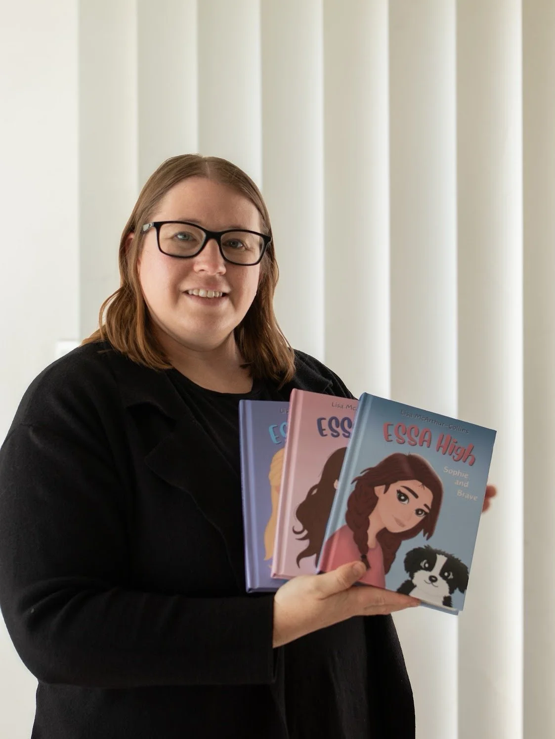 A woman with glasses and long brown hair smiling and holding three illustrated books titled 'ESSA High'.