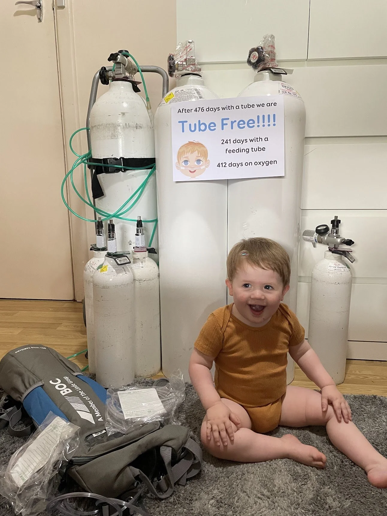 A young child sitting on a gray carpeted floor, smiling and laughing, surrounded by oxygen tanks and medical supplies. A sign on the tanks indicates the child has been free of a feeding tube for 241 days and on oxygen for 412 days.