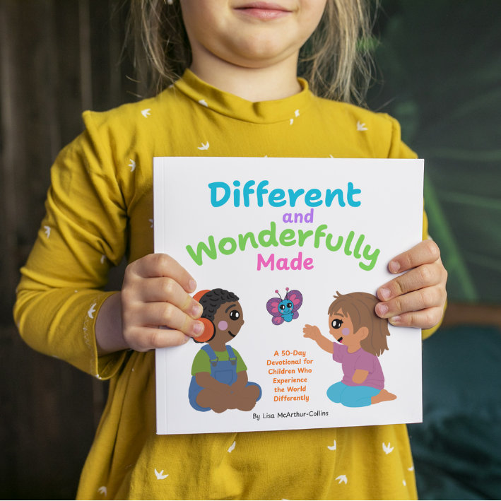 Child wearing a yellow shirt holding a book titled 'Different and Wonderfully Made' in a room with a wooden wall and a large green plant.