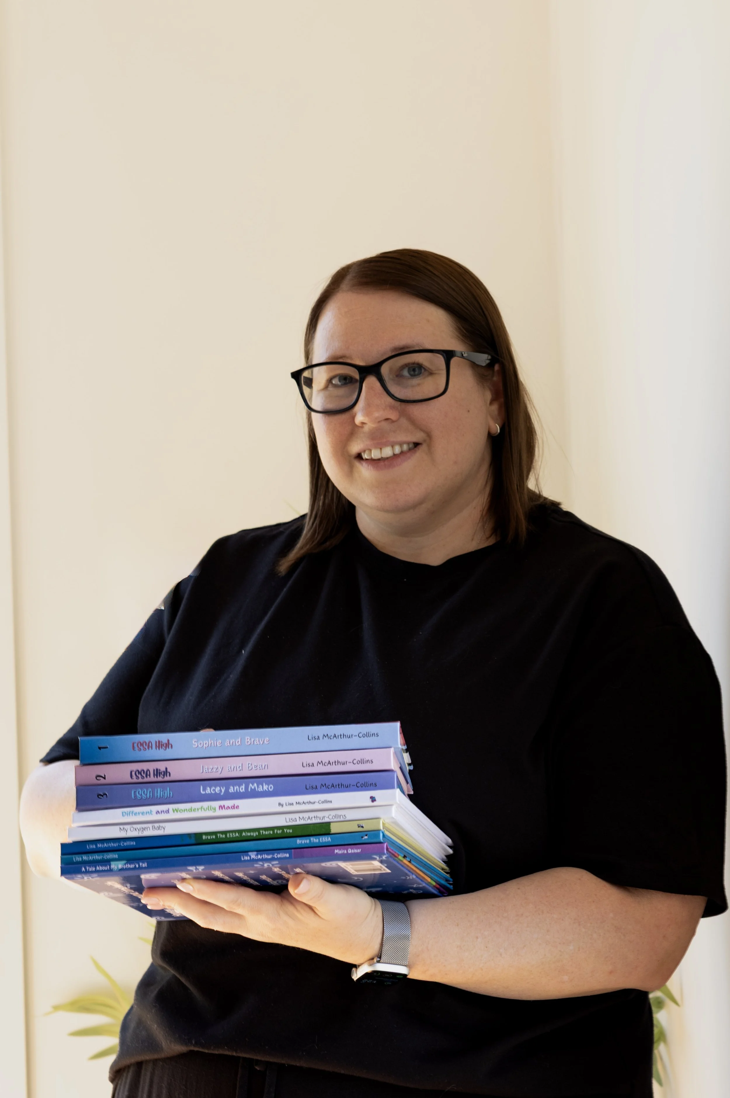 A woman with red hair, black glasses, and wearing a black shirt, holds a stack of colorful books related to ESSA at her chest. She is smiling and standing indoors against a light-colored wall.