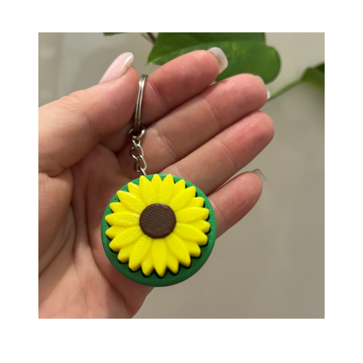 A hand holding a keychain with a sunflower charm. The sunflower has yellow petals and a brown center, and is set against a green background.