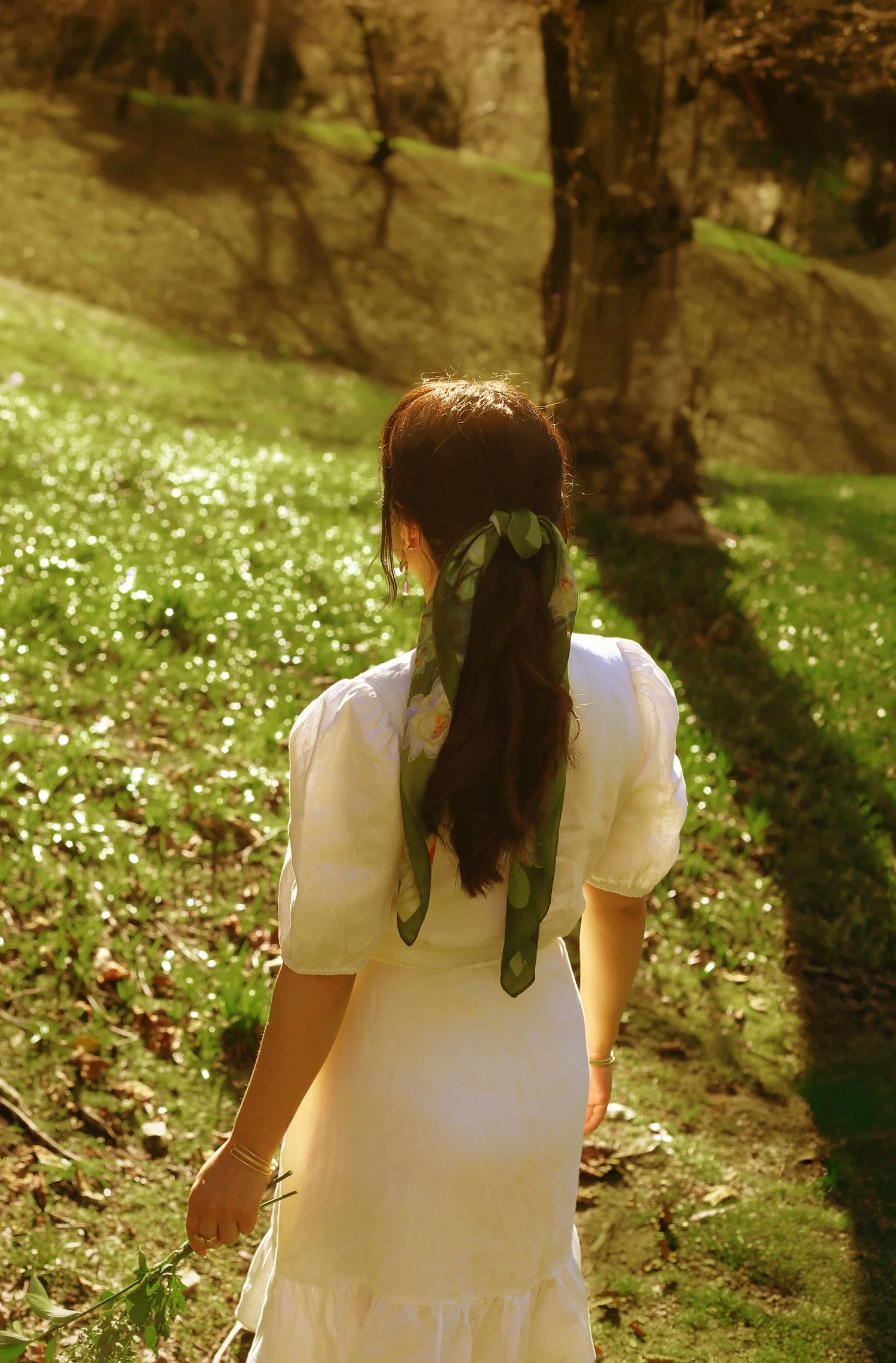 A woman is standing with her back to the camera in a sunlit forest, wearing a white dress and a green patterned scarf in her hair, holding a branch with leaves.