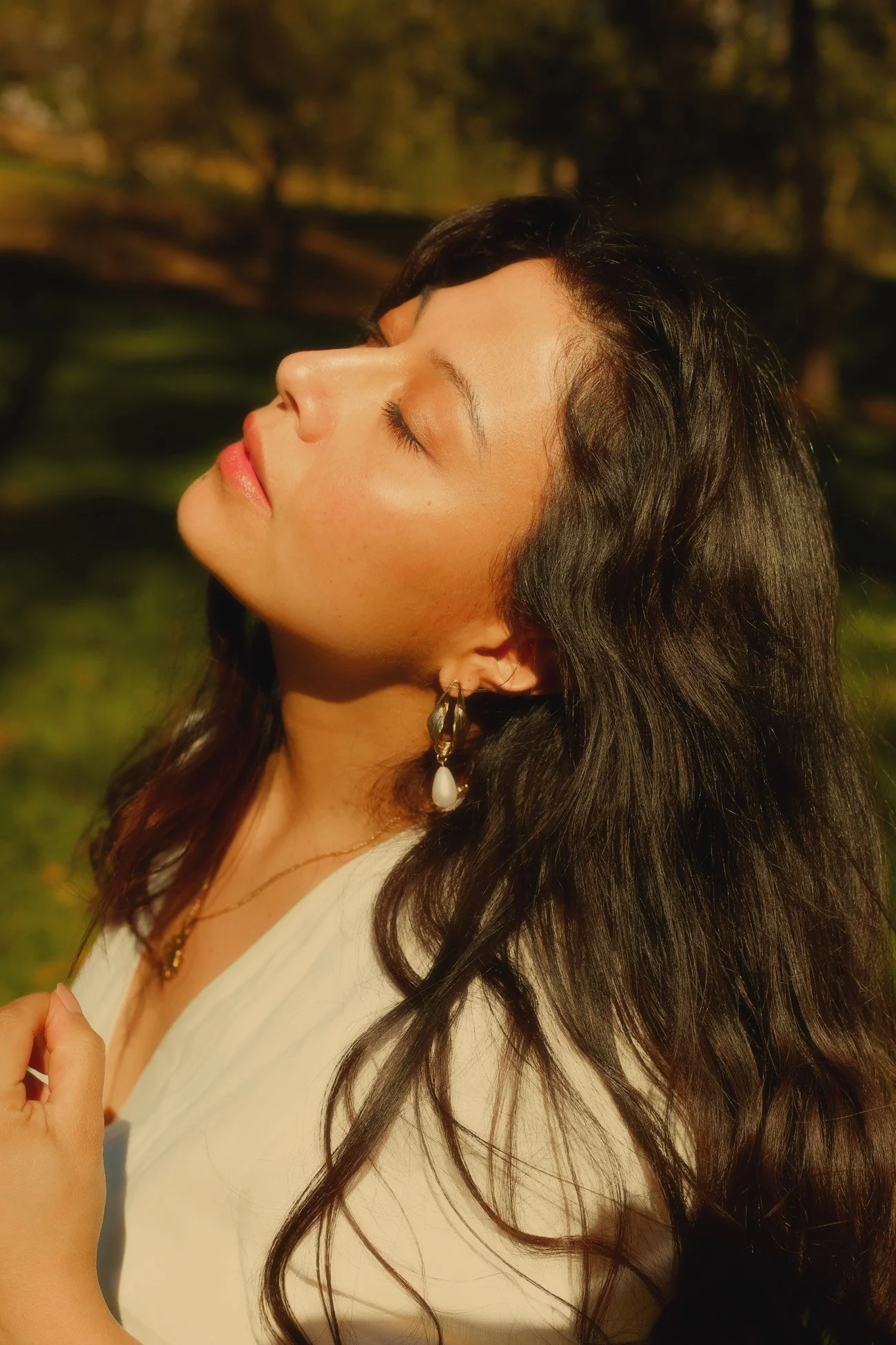 A woman with long dark wavy hair and earrings, eyes closed, enjoying sunlight outdoors with a blurred natural background.