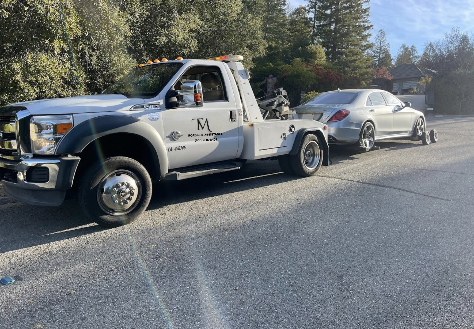 A tow truck is attached to the front of a silver Mercedes-Benz car on a suburban street. The tow truck is white with black text that reads 'Roadside Assistance' and a phone number. The street is lined with trees and a house can be seen in the background. The car's front left tire is damaged and on a wheel chock.