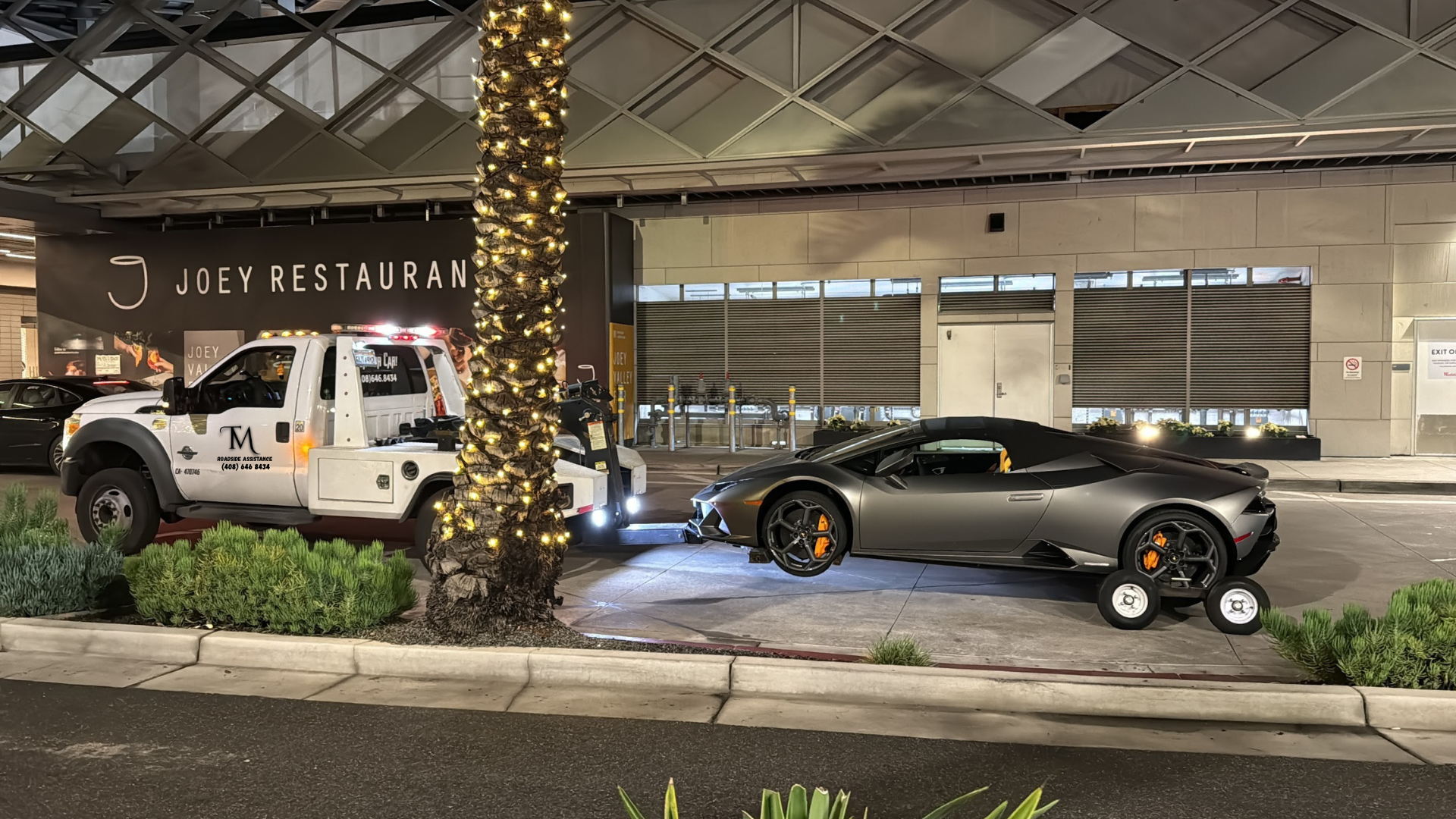 A silver Lamborghini convertible parked in front of Joey Restaurant, with a tree wrapped in lights and a white tow truck nearby. The scene takes place at night with some greenery in the foreground.