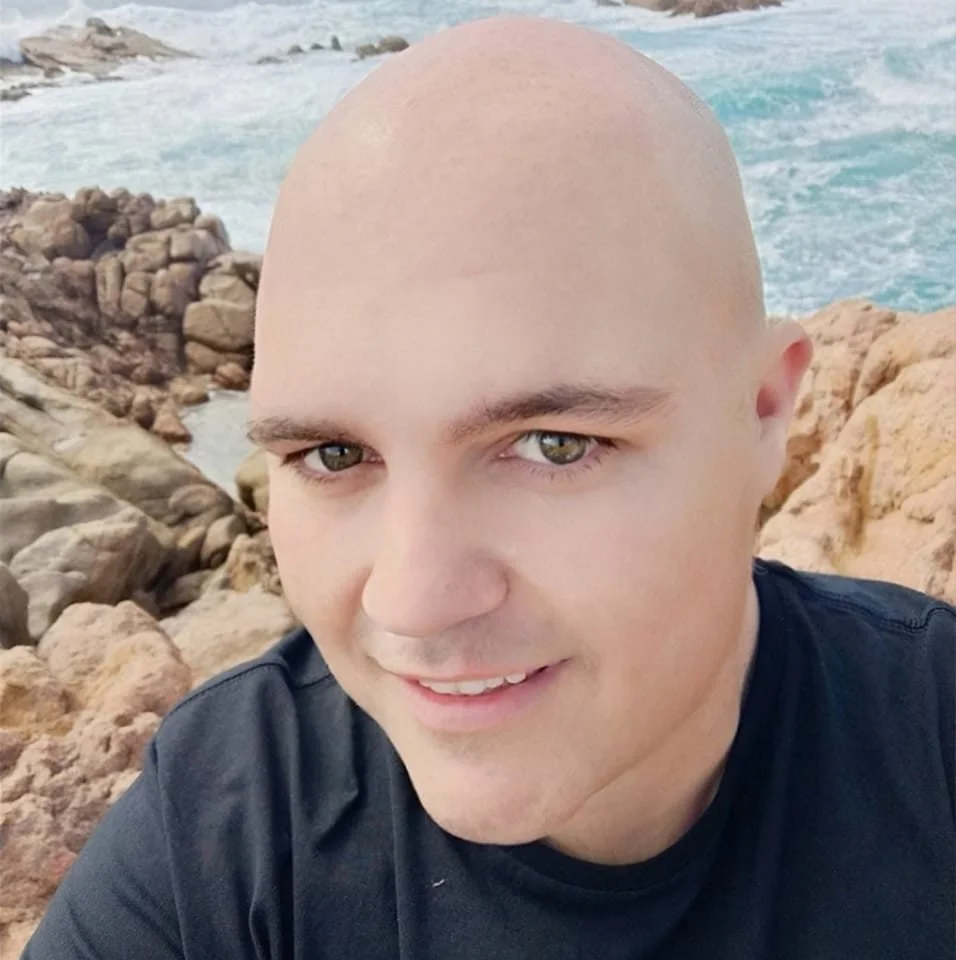 A man with a shaved head and light skin taking a selfie outdoors near rocky terrain and ocean waves.