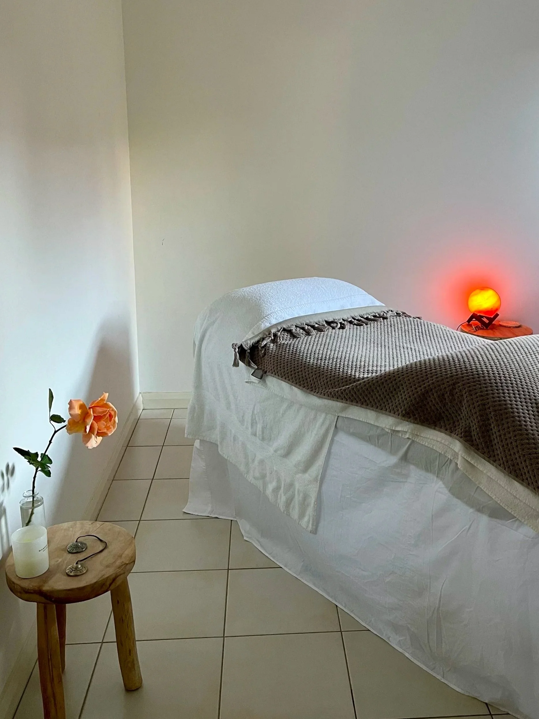 A bedroom scene with a bed covered in white and brown blankets, a small wooden side table with candles, stones, and a plant with peach-colored flowers, beige tiled floor, and a red-orange glowing lamp on a nightstand.