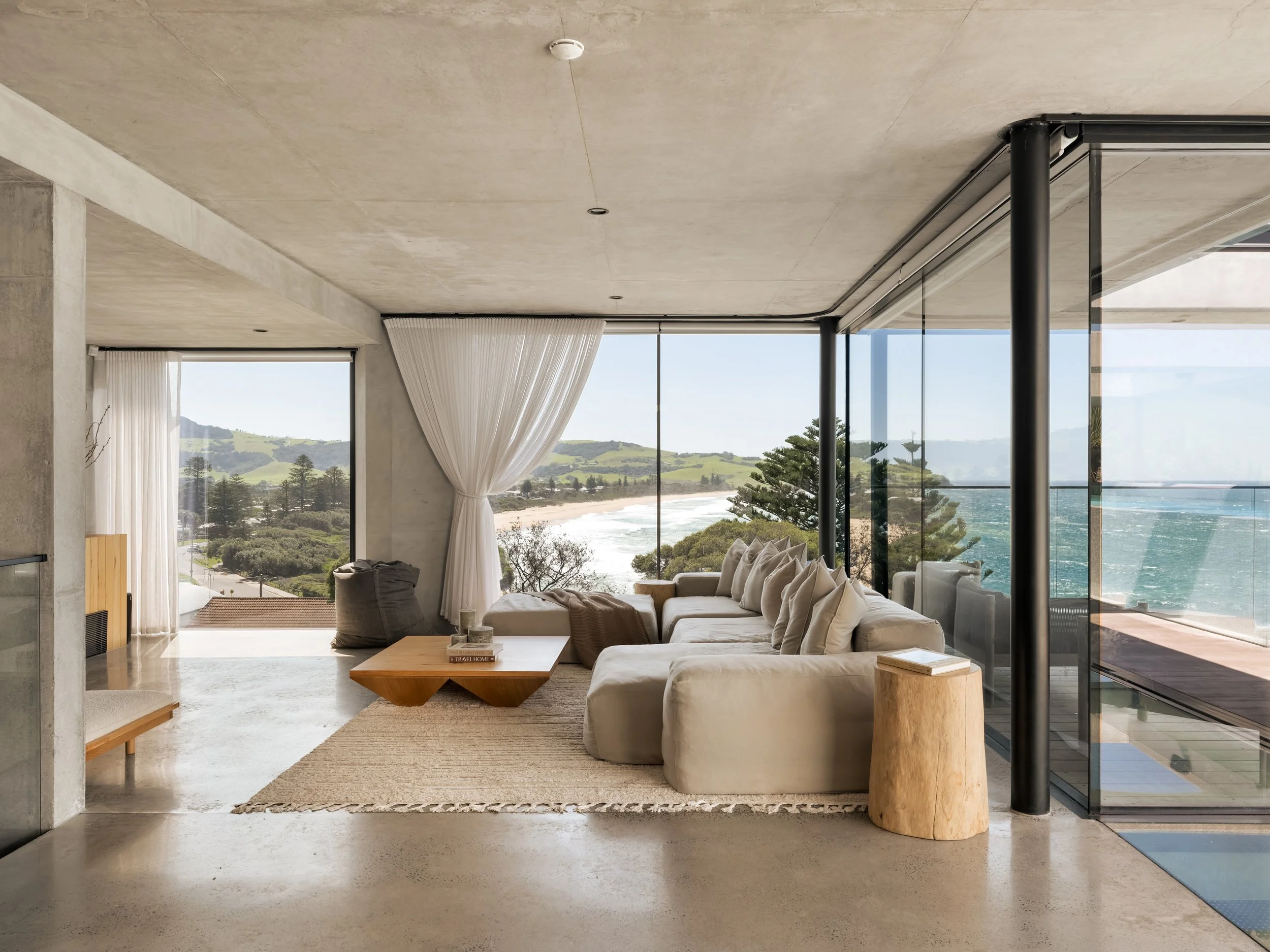 Modern living room with large glass windows showcasing ocean view, white sectional sofa, wooden coffee table, and minimal decor.