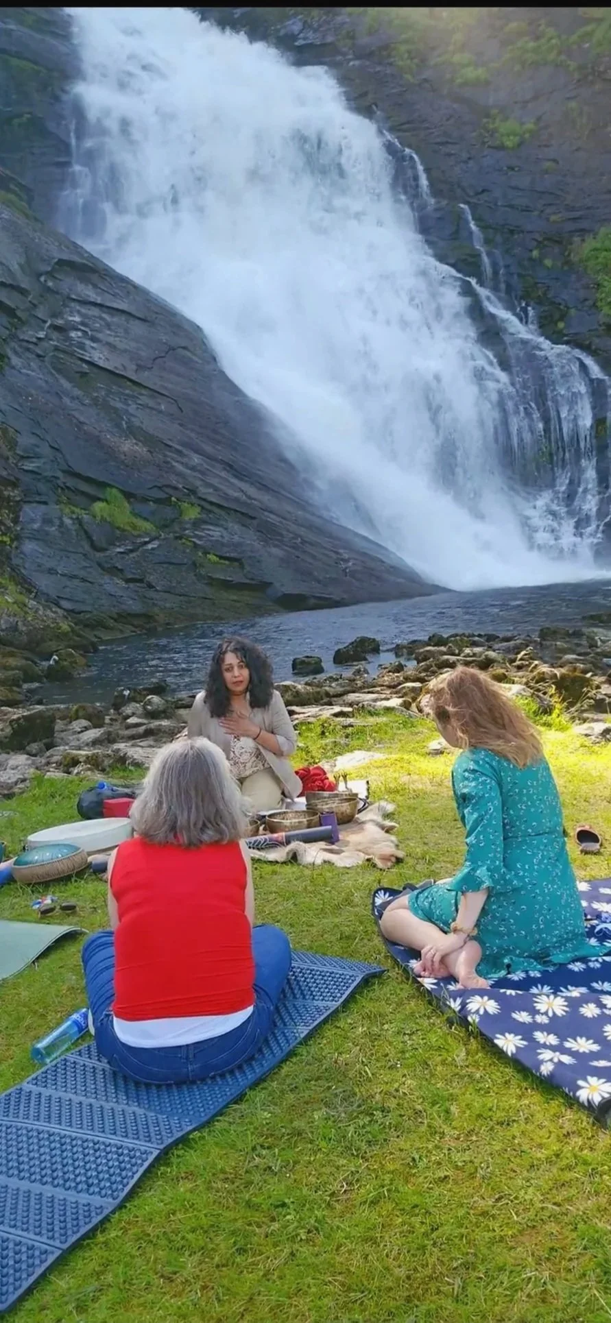 Three women sitting on blankets and mats on grass near a rocky riverbank with a large waterfall in the background, engaged in a spiritual or meditative activity.