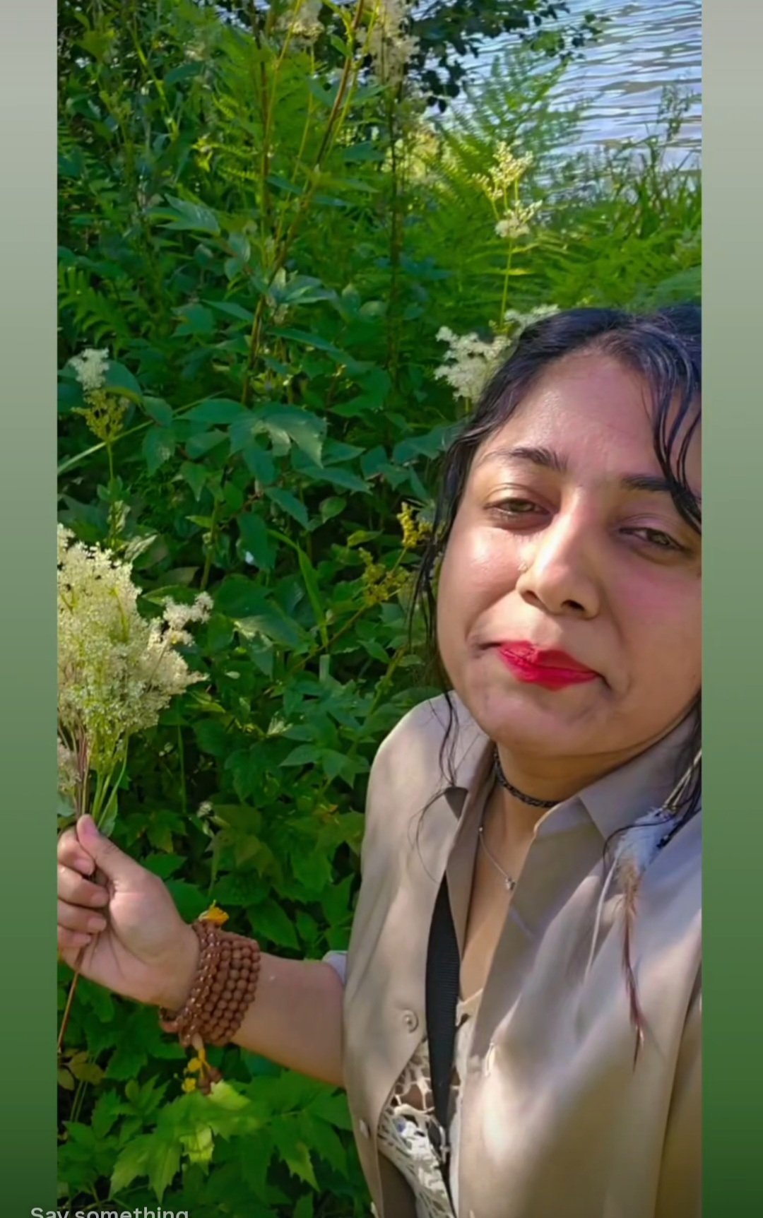 A woman taking a selfie outdoors, holding a bunch of white flowers, with green foliage and a body of water in the background.