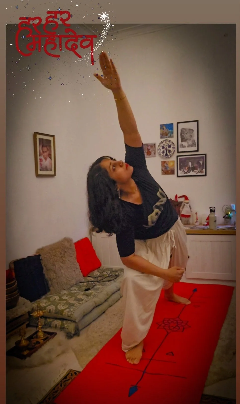 A woman practicing yoga on a red mat in a living room, with one arm raised and she is balancing on one leg, looking upward. The room has framed pictures on the wall and a couch with cushions and a rug on the floor.